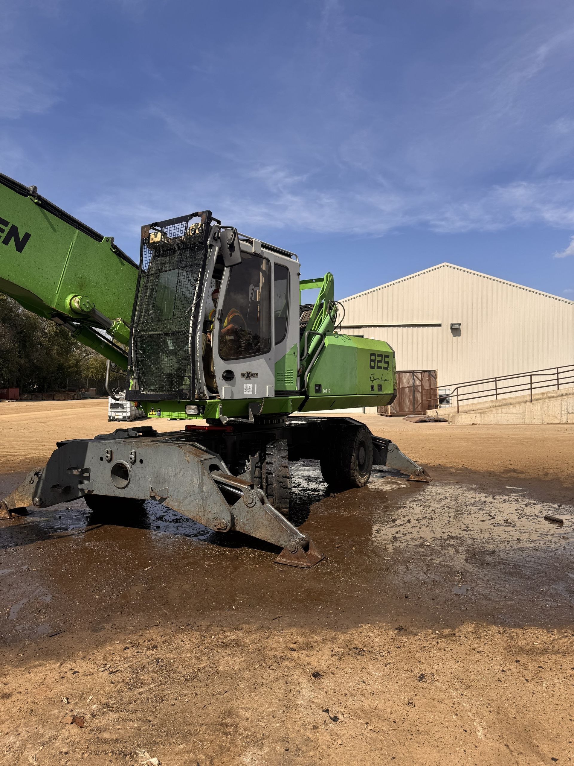 Damaged green construction machine with crushed front end in a muddy yard beside a beige building.