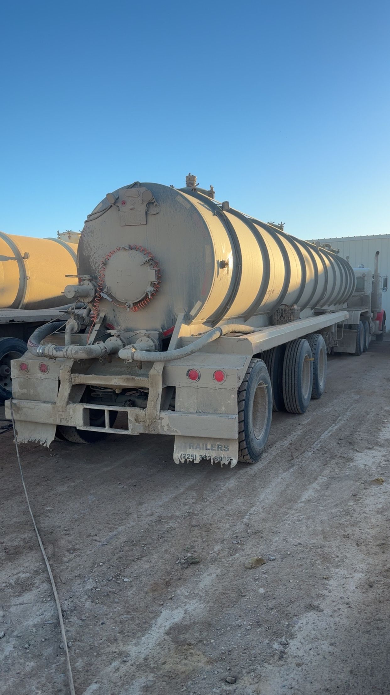 Large yellow industrial tank trailer parked on a muddy dirt lot under a clear blue sky.