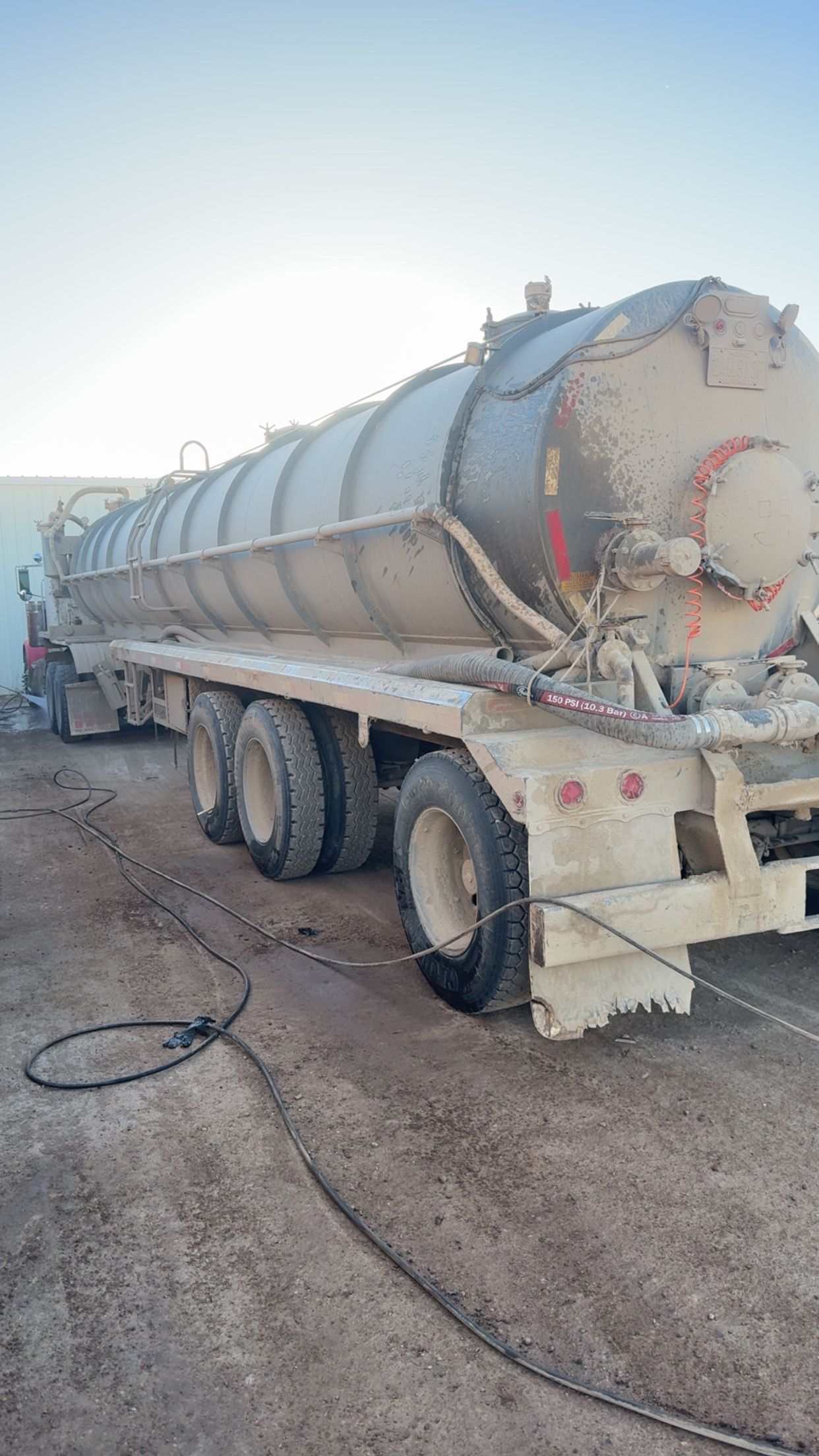 Dusty white tanker truck parked on a gravel lot under a bright sky