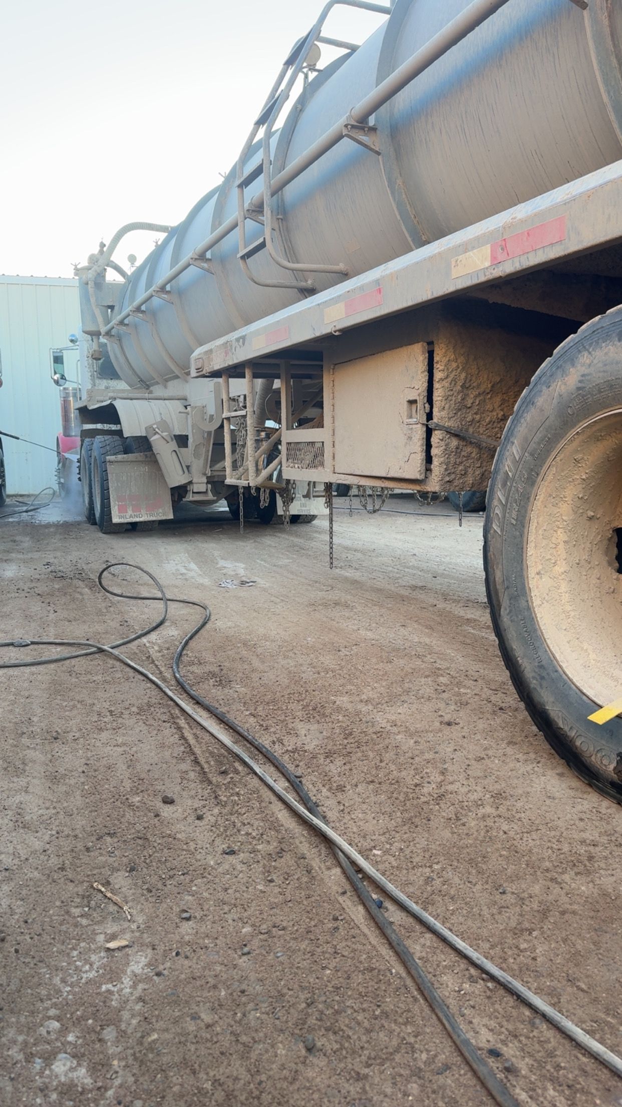 Large tanker truck parked on a dirt lot, side view with hoses on the ground and a large wheel visible.