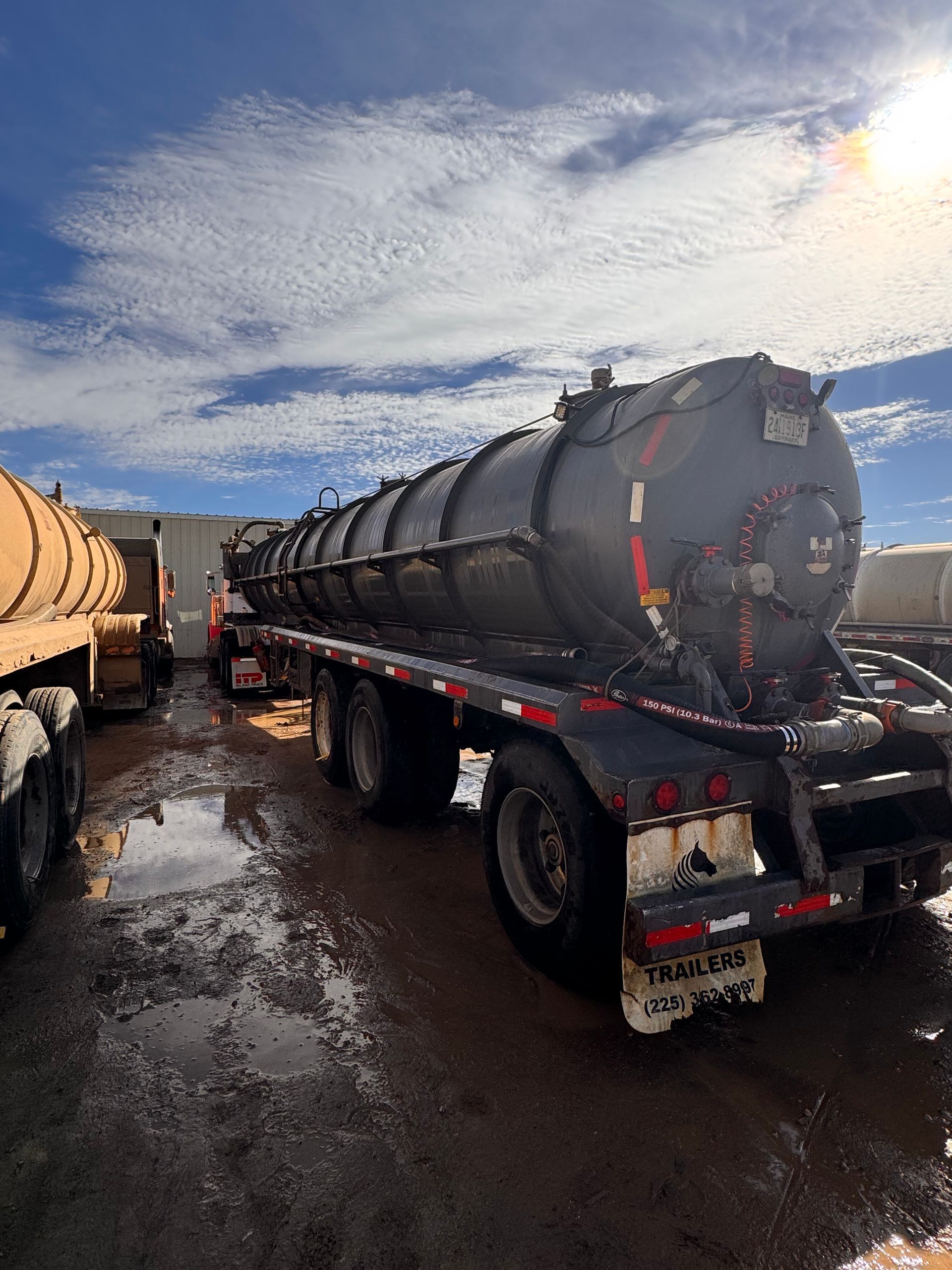 Black tanker truck parked in a wet lot under a cloudy sky, with another tanker partly visible beside it