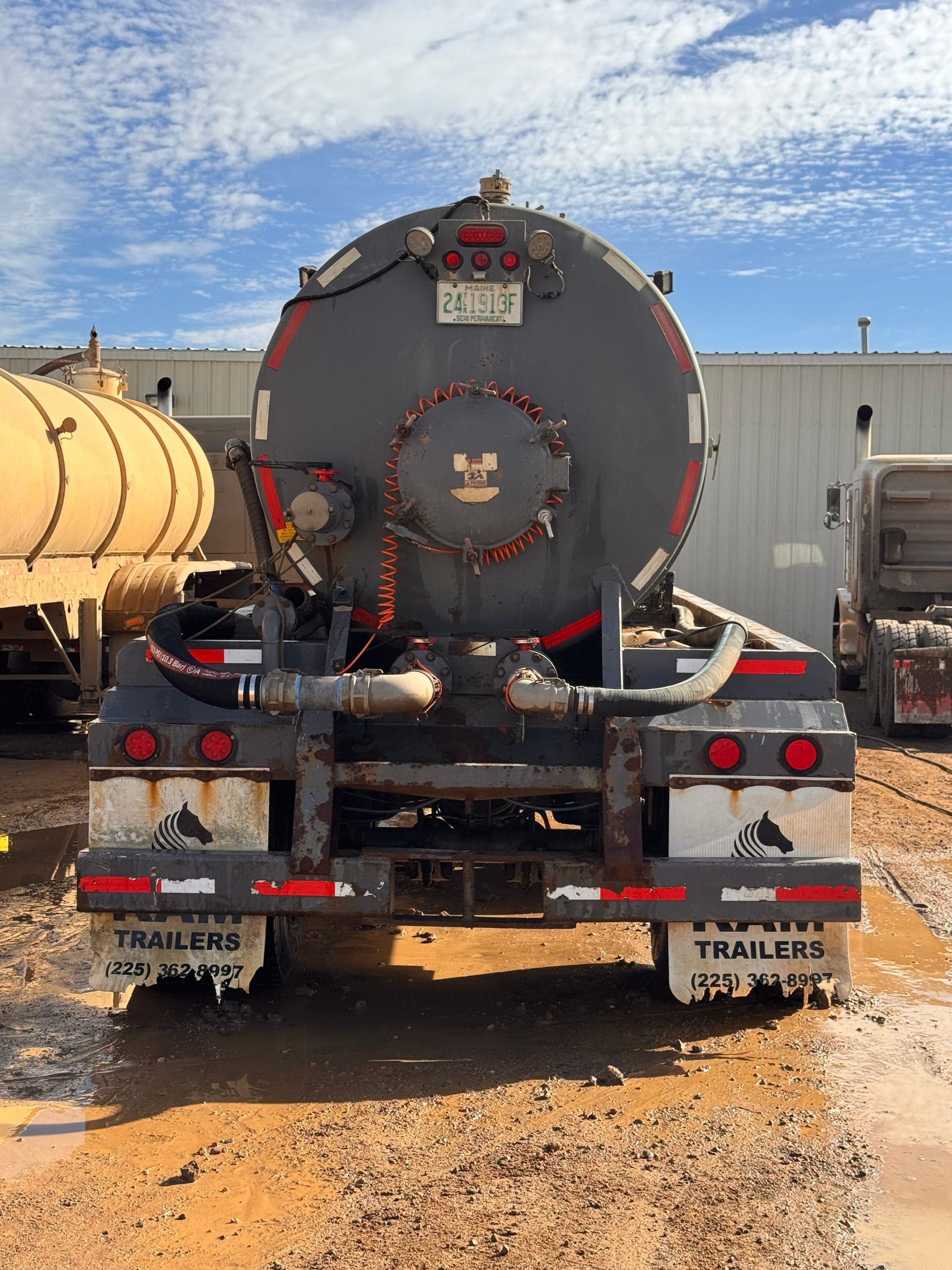 Rear view of a gray tanker truck with red lights parked on a muddy lot under a cloudy sky