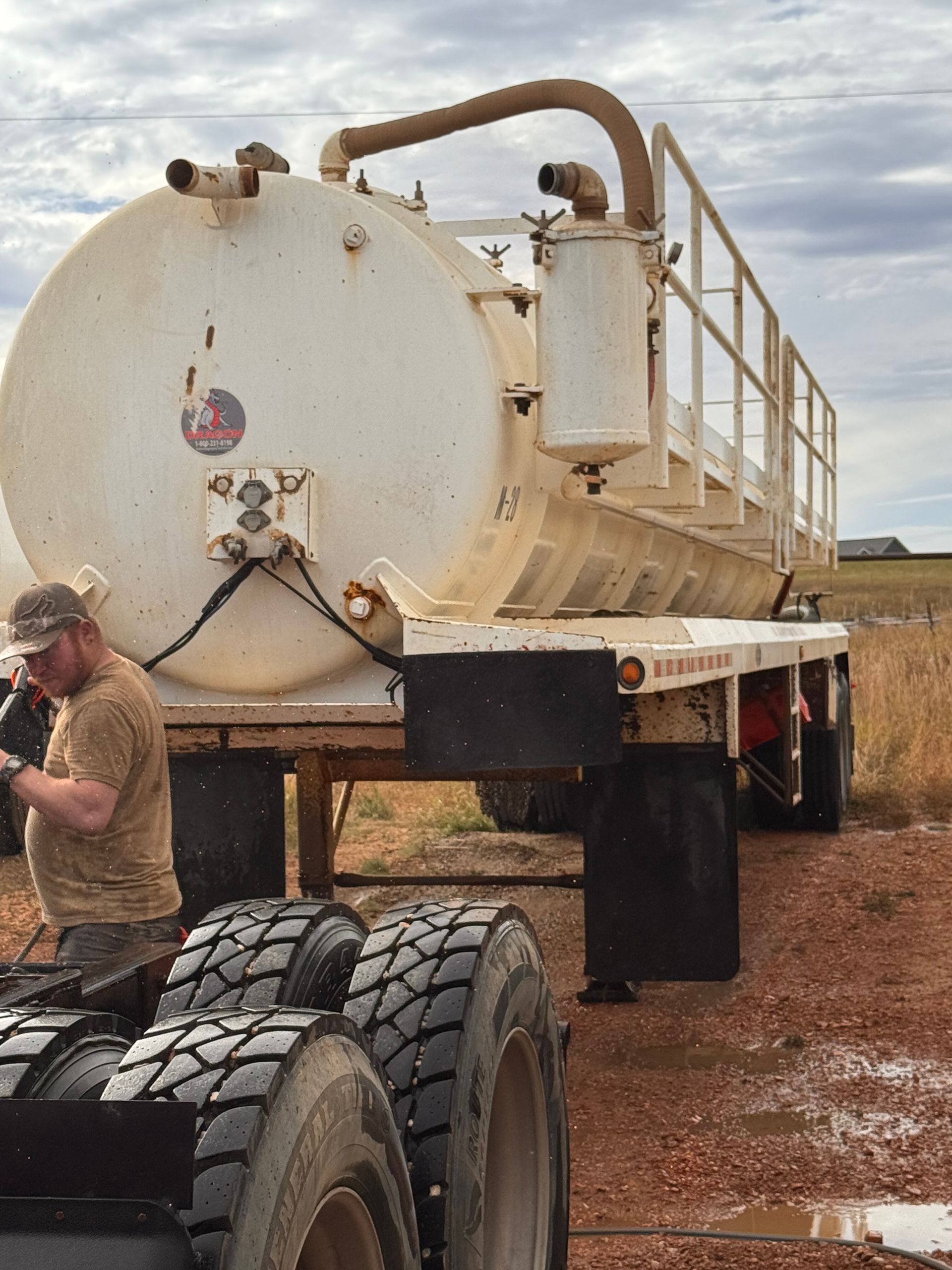 Large white tanker trailer with oversized tires in a muddy field, beside a person in a cap