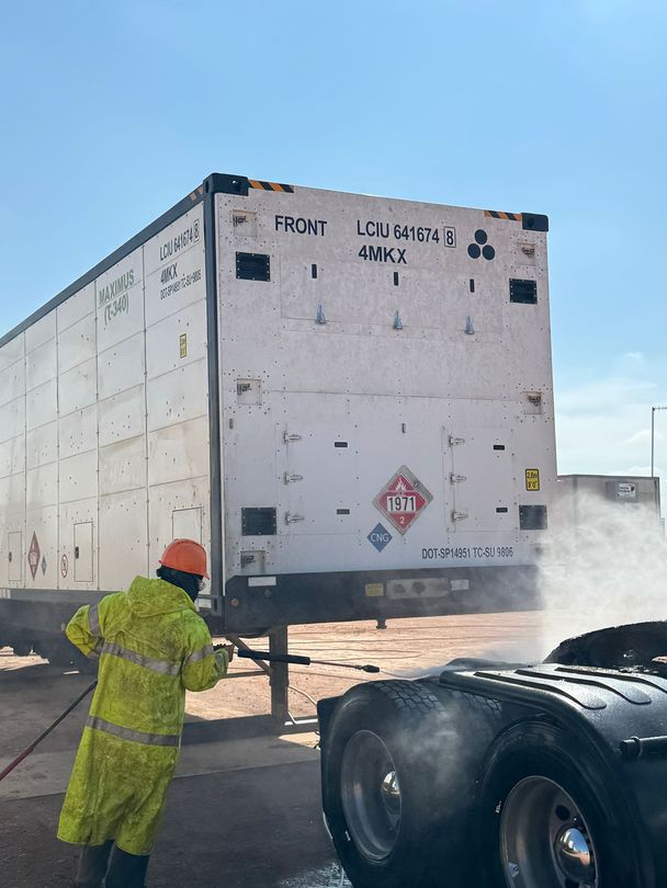 Worker in yellow safety gear inspects a cargo container beside a truck, with steam or dust rising nearby.