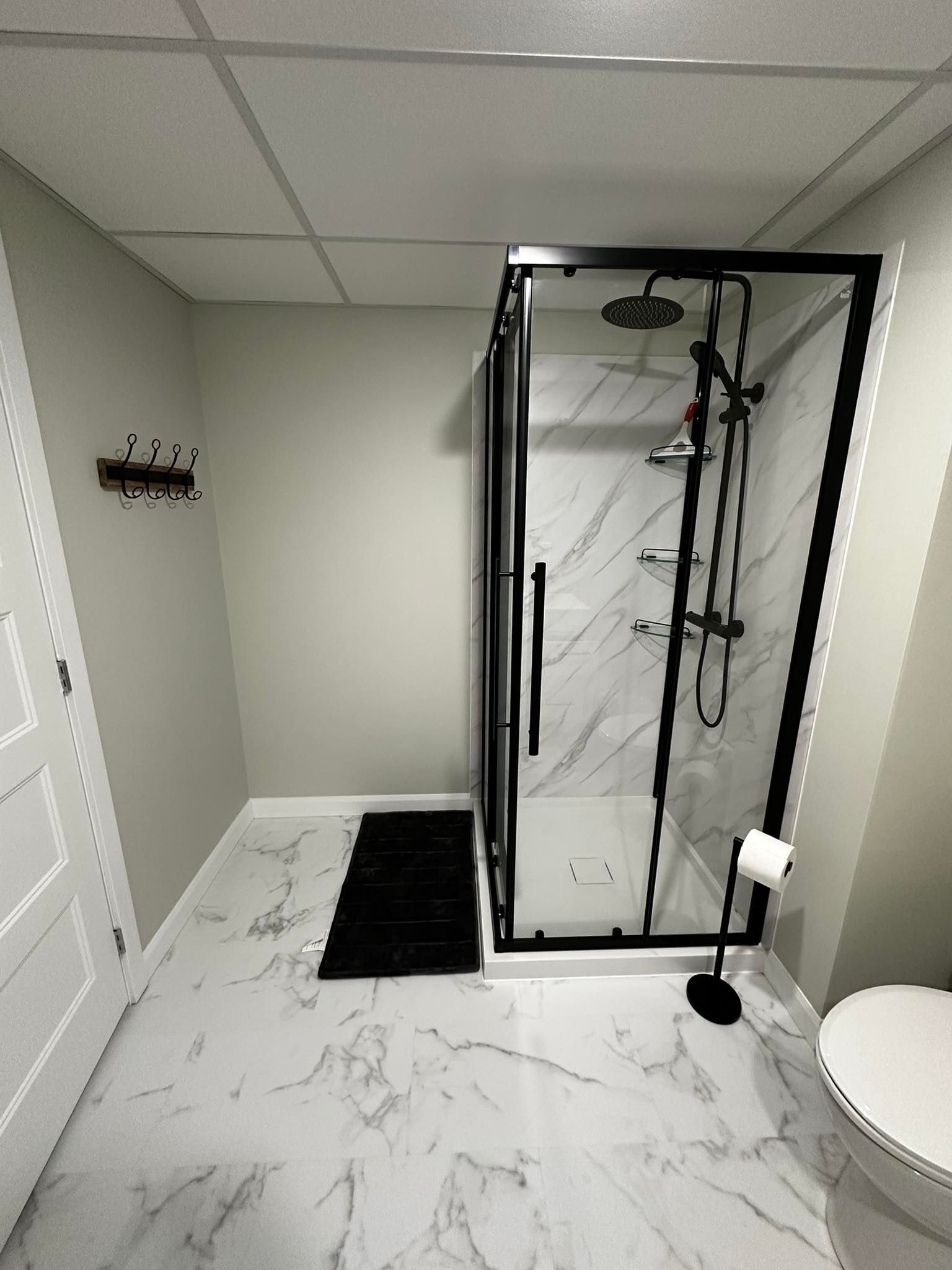 Salle de bains moderne avec douche en verre, sol imitation marbre, tapis noir et toilettes d'angle.