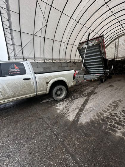 Une camionnette blanche a reculé dans une remorque benne noire à l'intérieur d'un grand hangar au toit incurvé, avec des débris au sol.
