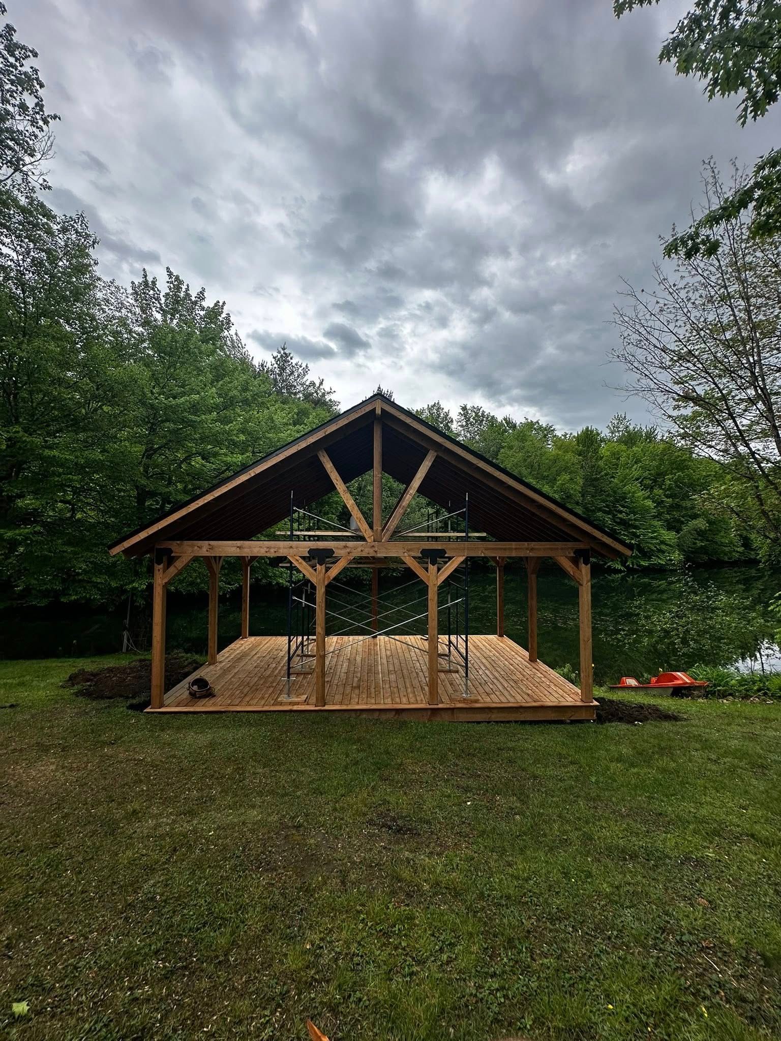 Pavillon en bois dans un champ herbeux sous un ciel nuageux, avec des bottes de foin à l'intérieur et des arbres derrière.