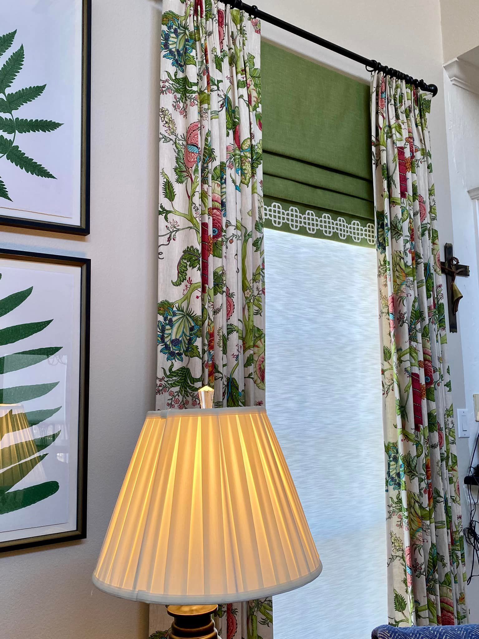 Floral curtains frame a window with a green shade; a lamp with a pleated shade is in the foreground.