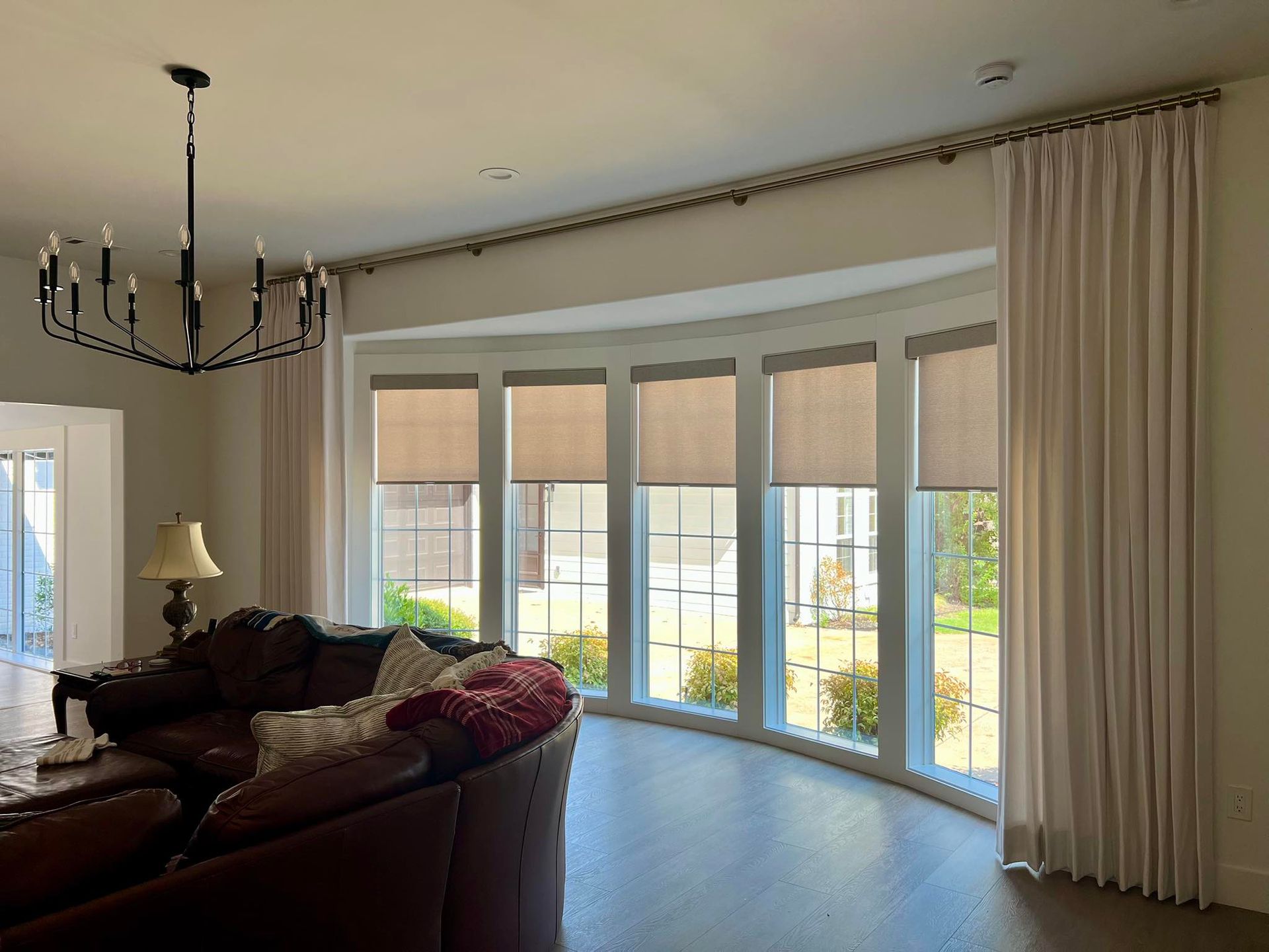 Living room with large curved windows, beige drapes, and a chandelier over a leather sofa.