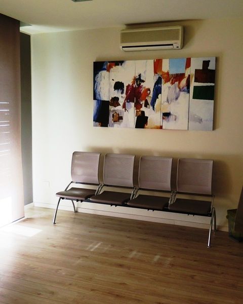 Sala d'attesa moderna con quattro sedie grigie, un quadro astratto alle pareti, pavimento in legno e un condizionatore d'aria sopra.
