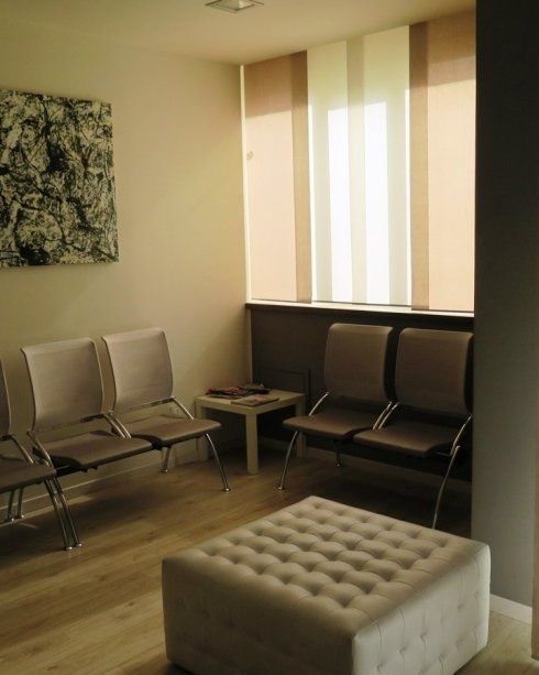 Piccola sala d'attesa con sedie in metallo, un tavolino basso, un pouf imbottito e alte tende alle finestre.