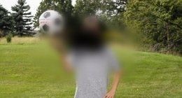 Figura sfocata in piedi su un campo erboso che tiene in mano un pallone da calcio bianco, con alberi sullo sfondo.