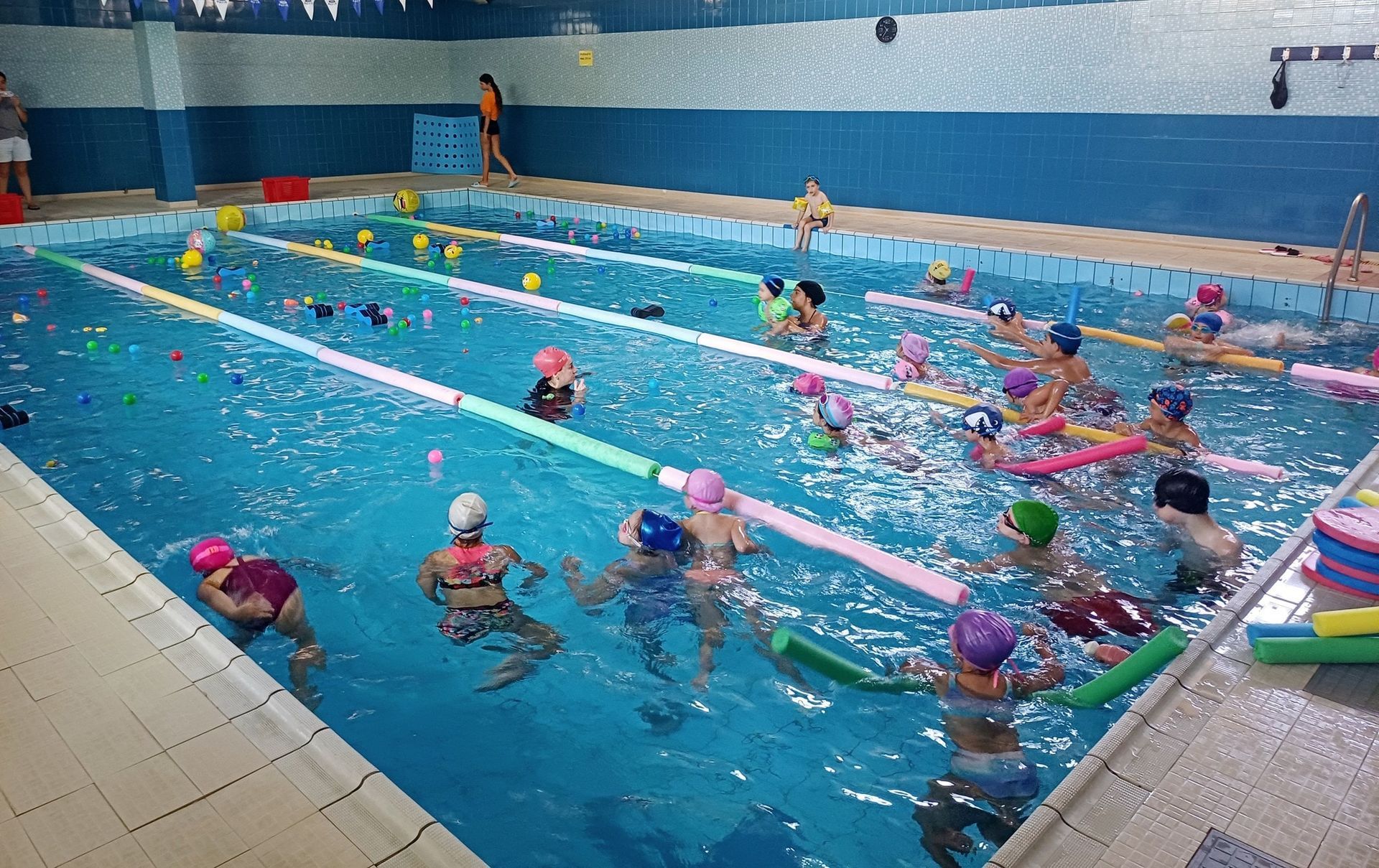 Bambini in piscina con istruttori di nuoto. Acqua azzurra, cuffie multicolori e galleggianti di gommapiuma.
