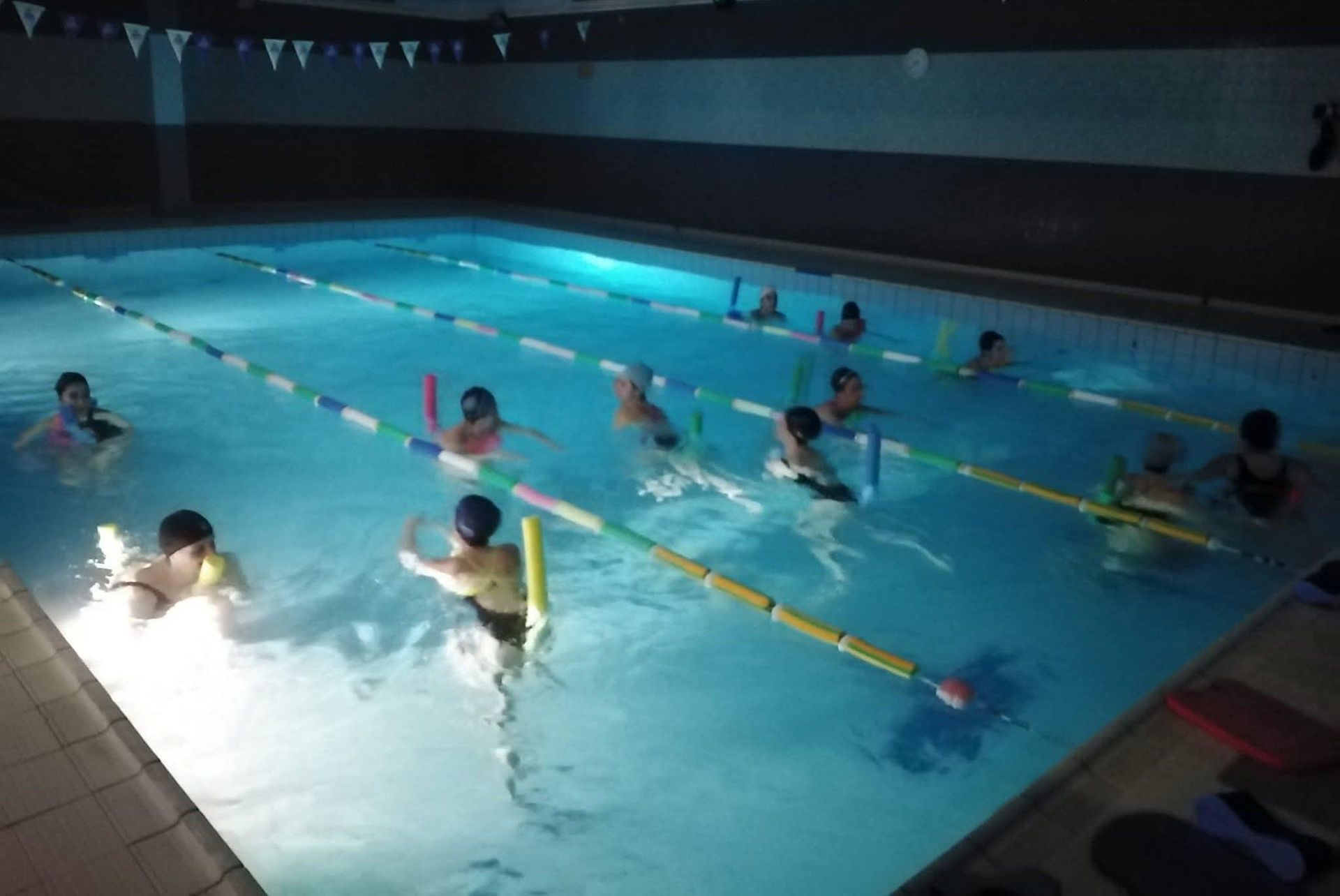 Bambini in piscina che imparano a nuotare, usando tubi di gommapiuma e bracciali galleggianti. Ambiente al chiuso.