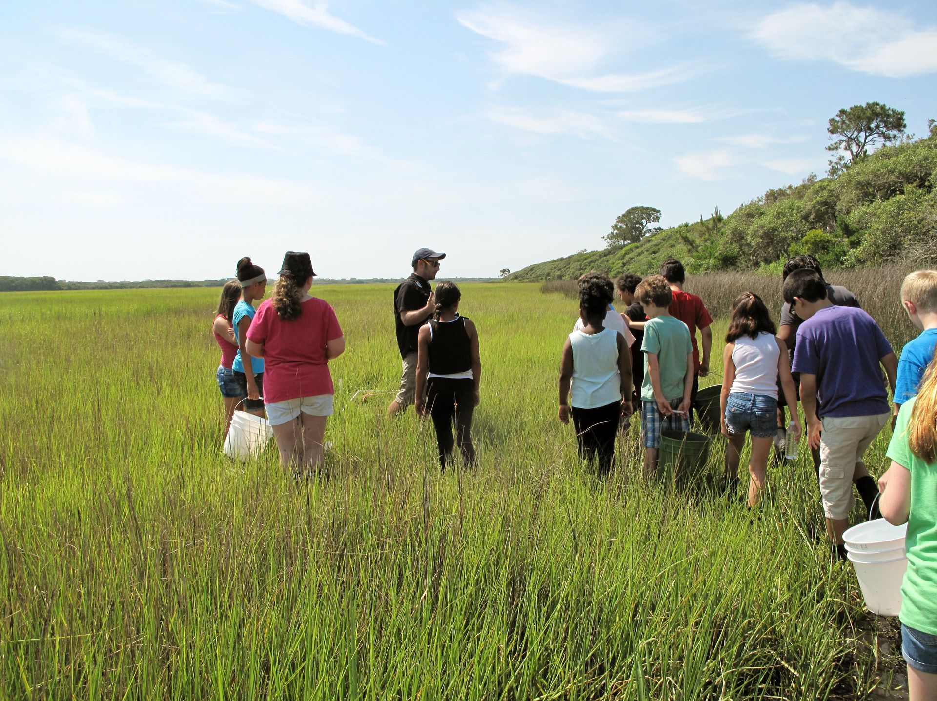 Group of people walking through a grassy field on a sunny day, with hills and blue sky in the background.