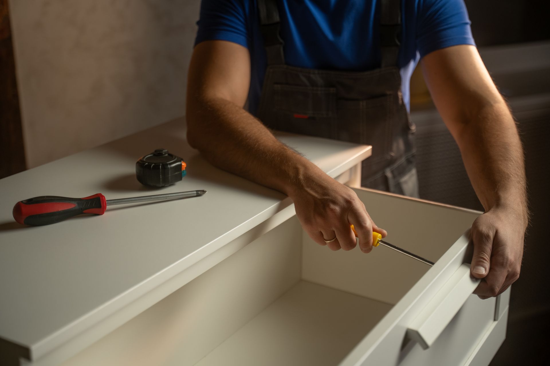 Man using a screwdriver to assemble a white cabinet drawer. A measuring tape and screwdriver are on the cabinet.