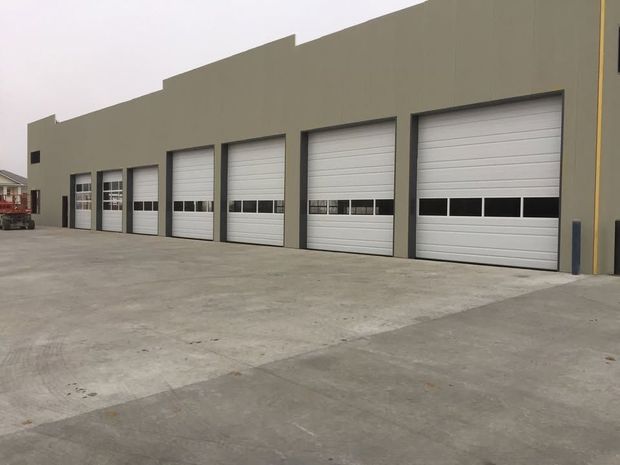 Row of white roll-up garage doors on a beige industrial building with a large concrete driveway