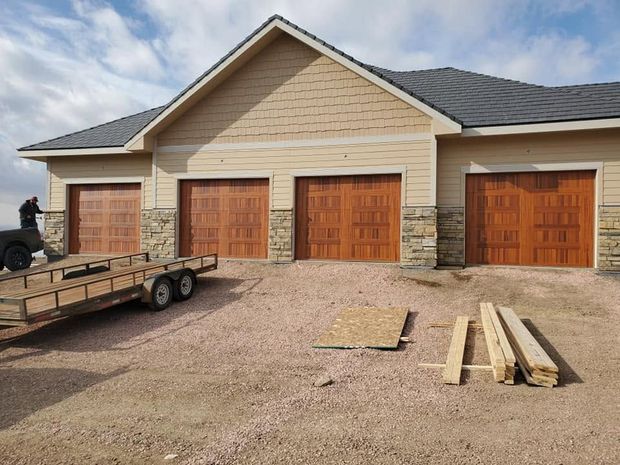 New garage with four wooden doors, gravel driveway, and lumber stacked in front under a cloudy sky
