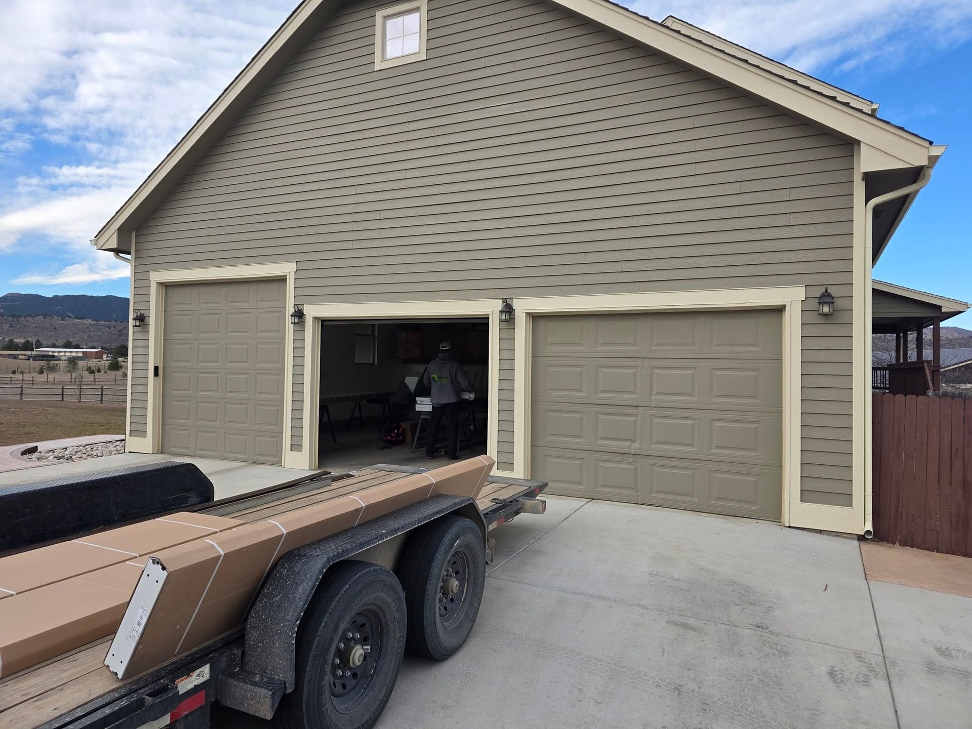 Tan house with three garage doors; trailer parked in driveway under a cloudy sky.