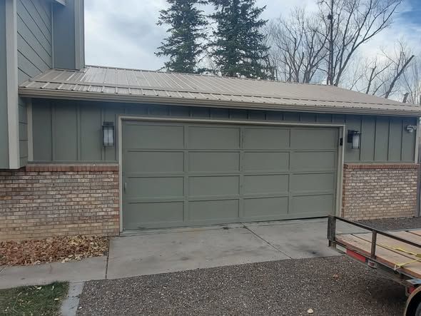 Closed green garage door beside brick walls on a driveway, with a trailer at right.
