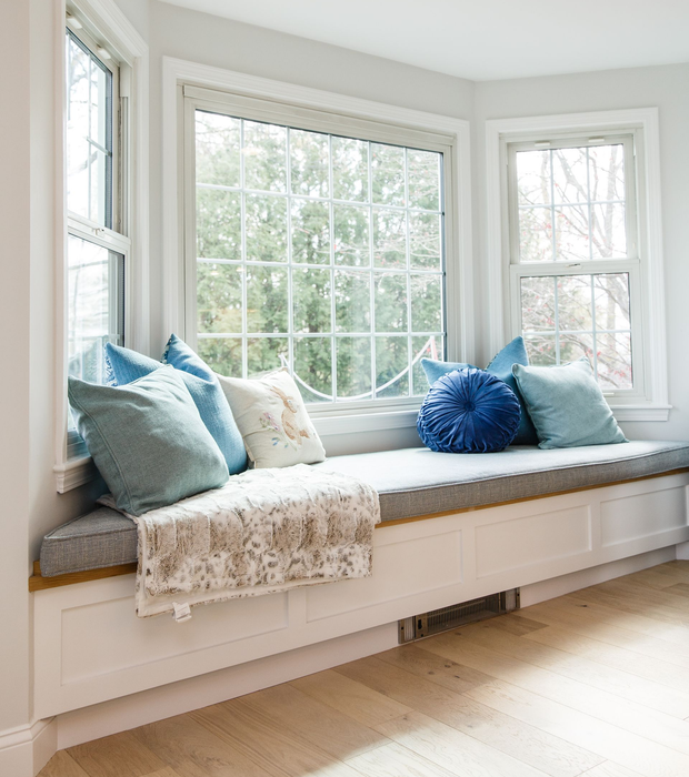 Bright bay window seat with gray cushion, blue pillows, and a soft blanket in a sunlit room
