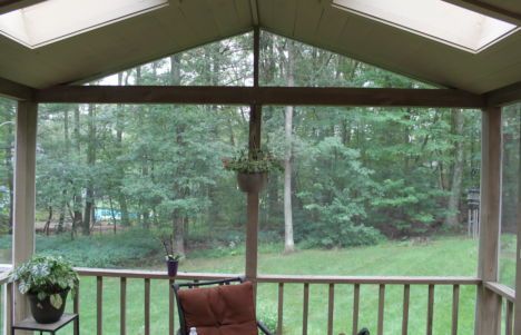 Covered porch overlooking a green wooded yard, with railing, hanging plant, and cushioned chair.