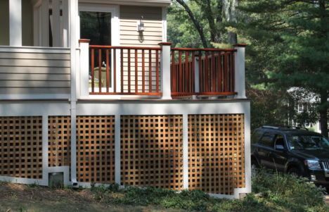 House porch with brown railing and lattice skirting beside a wooded driveway