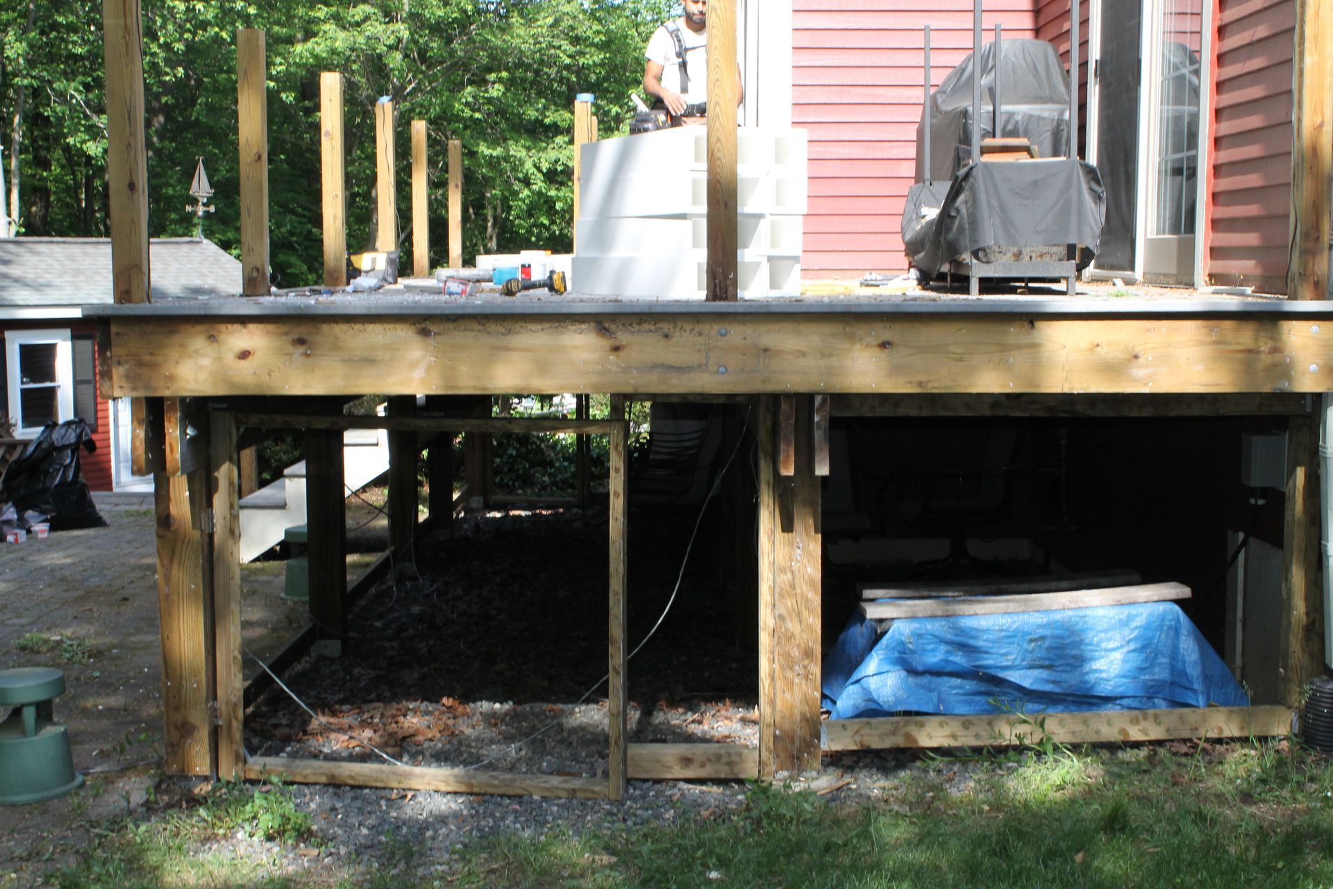 Wooden deck under construction beside a house, with posts, beams, stairs, and stored materials underneath