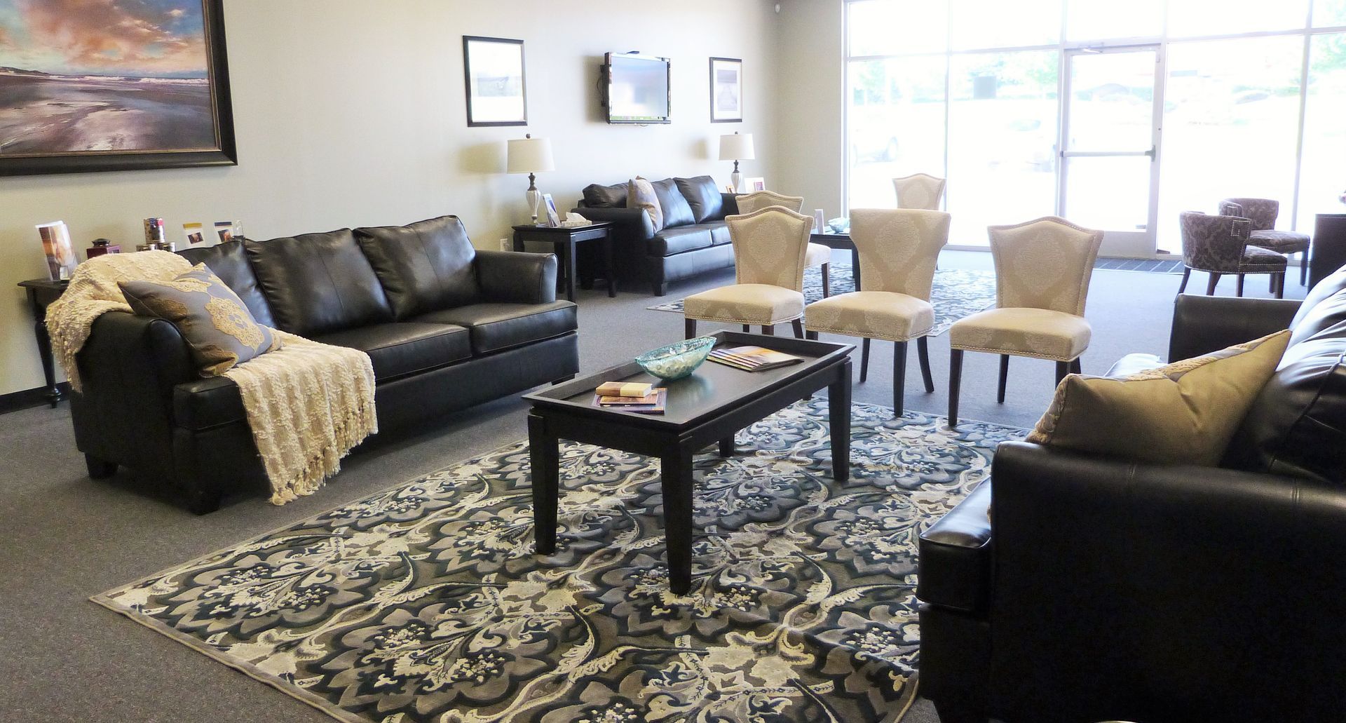 Modern living room with black sofas, patterned rug, coffee table, and beige chairs near bright windows.