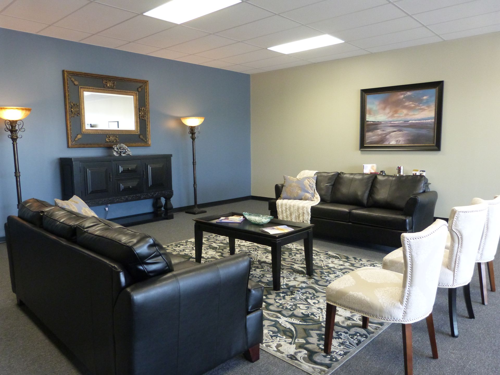 Modern living room with black leather sofas, patterned rug, TV, and white chairs under soft lighting