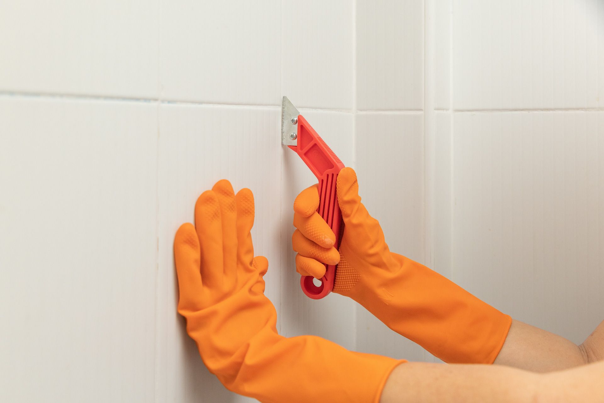 A person wearing orange rubber gloves is cleaning a tile wall with a spatula.