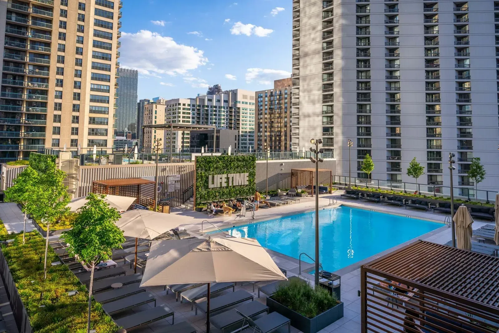 A rooftop pool area with lounge chairs, umbrellas, and trees, surrounded by high-rise buildings.