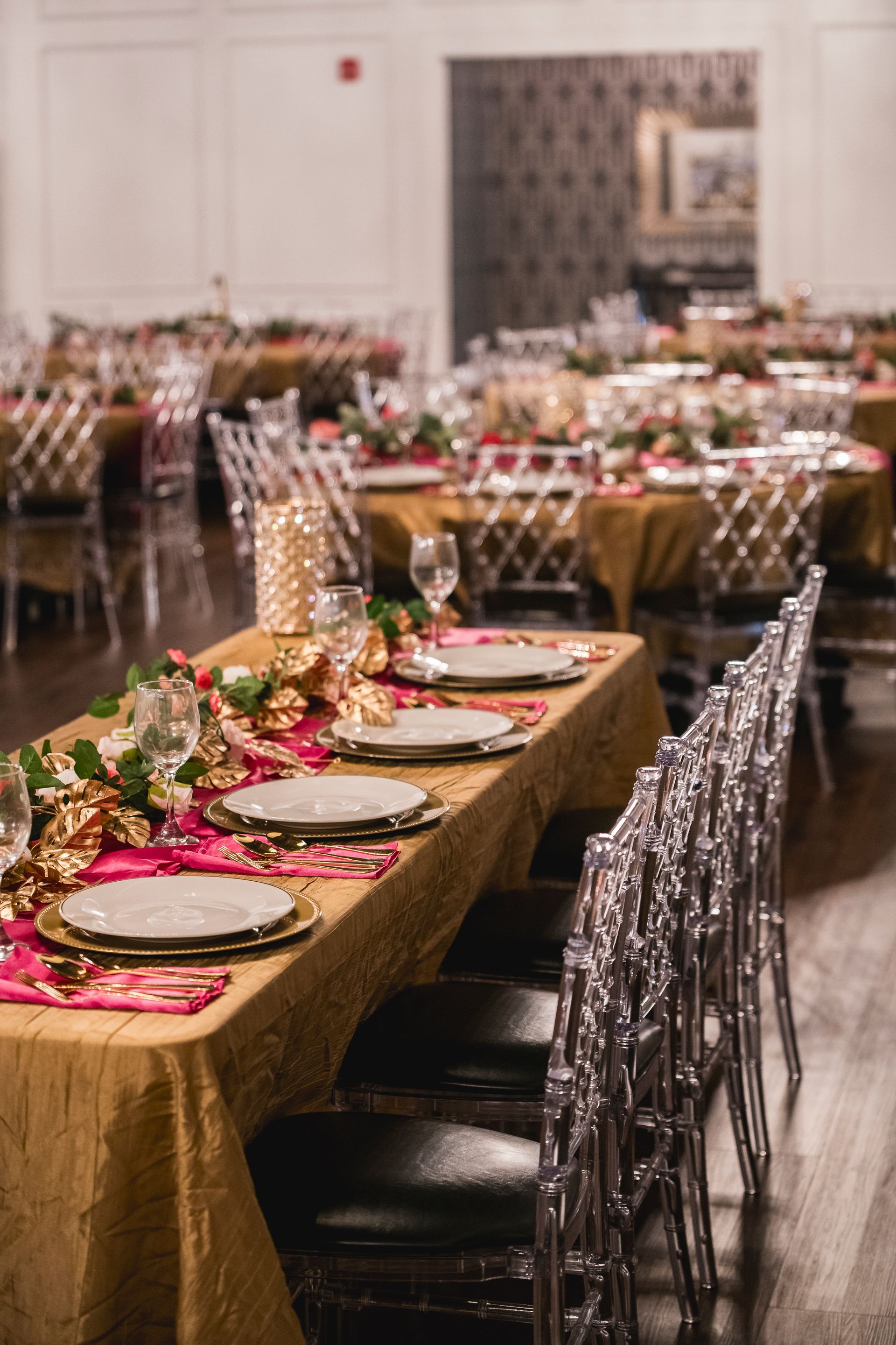 Elegant banquet table set with gold runner, floral centerpieces, and clear chairs in a decorated dining room
