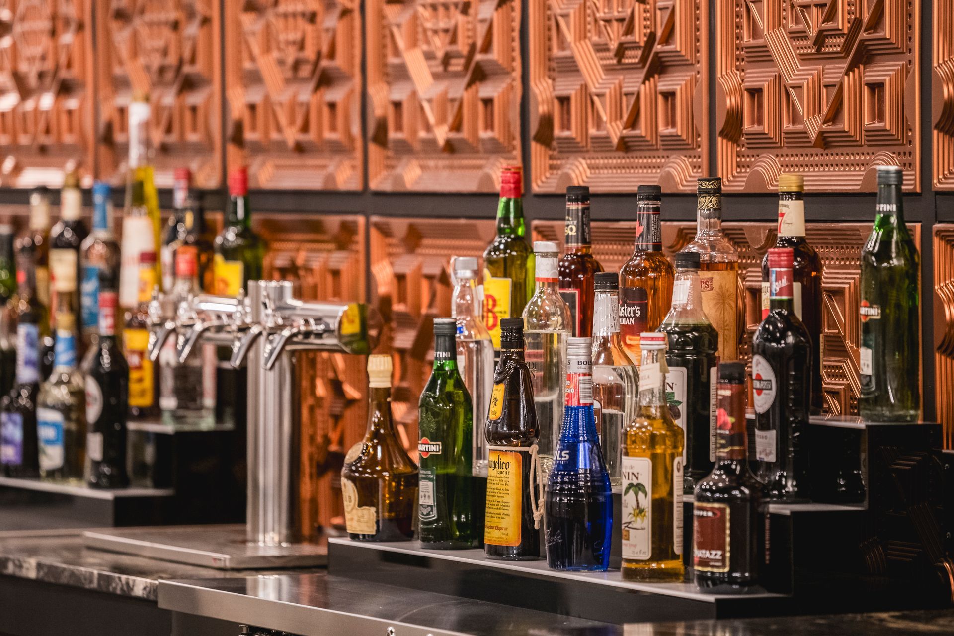 Bar counter lined with assorted liquor bottles and taps against an orange patterned wall.