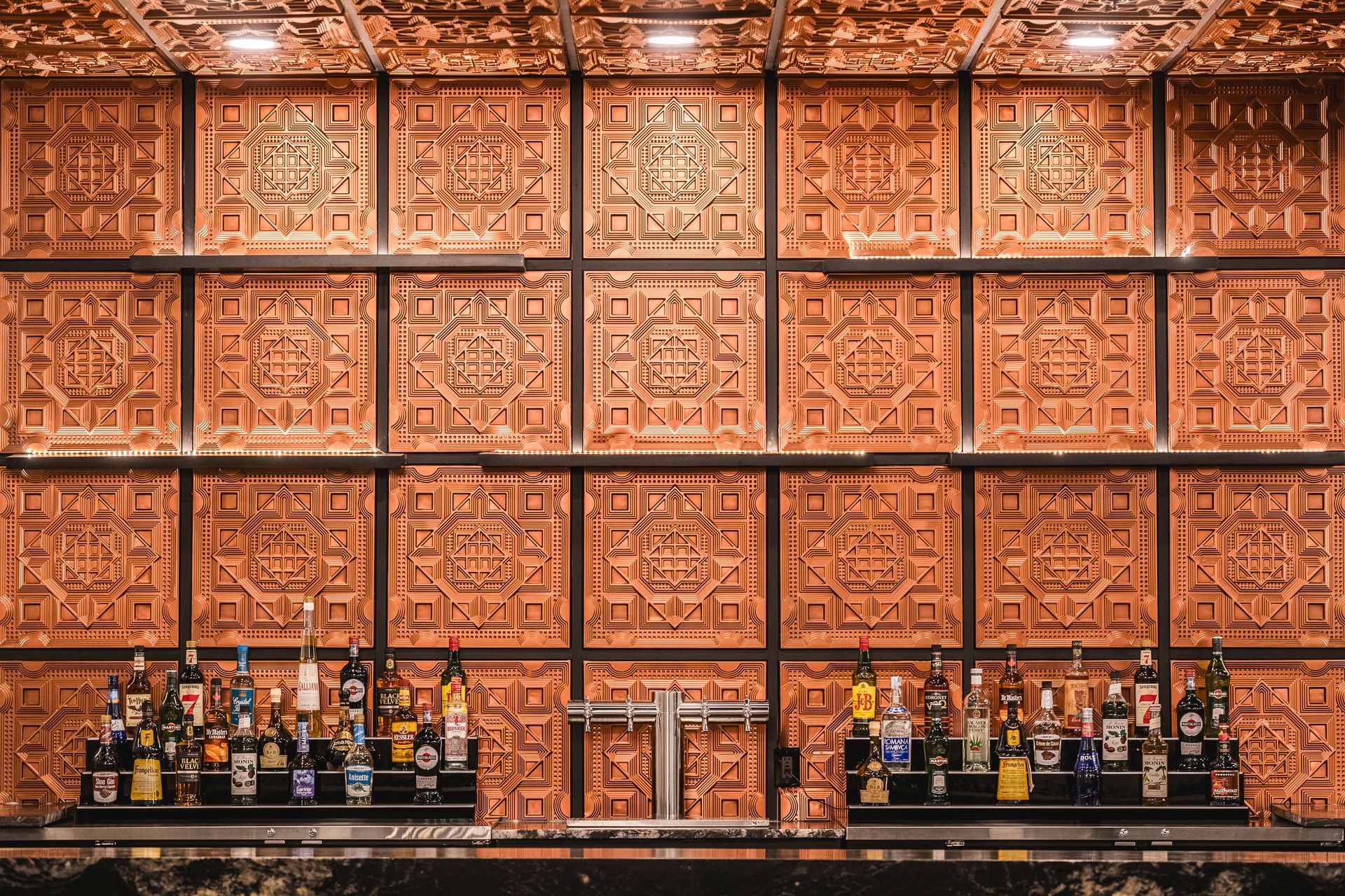 Rows of small bottles on a black counter before a copper-patterned wall backdrop