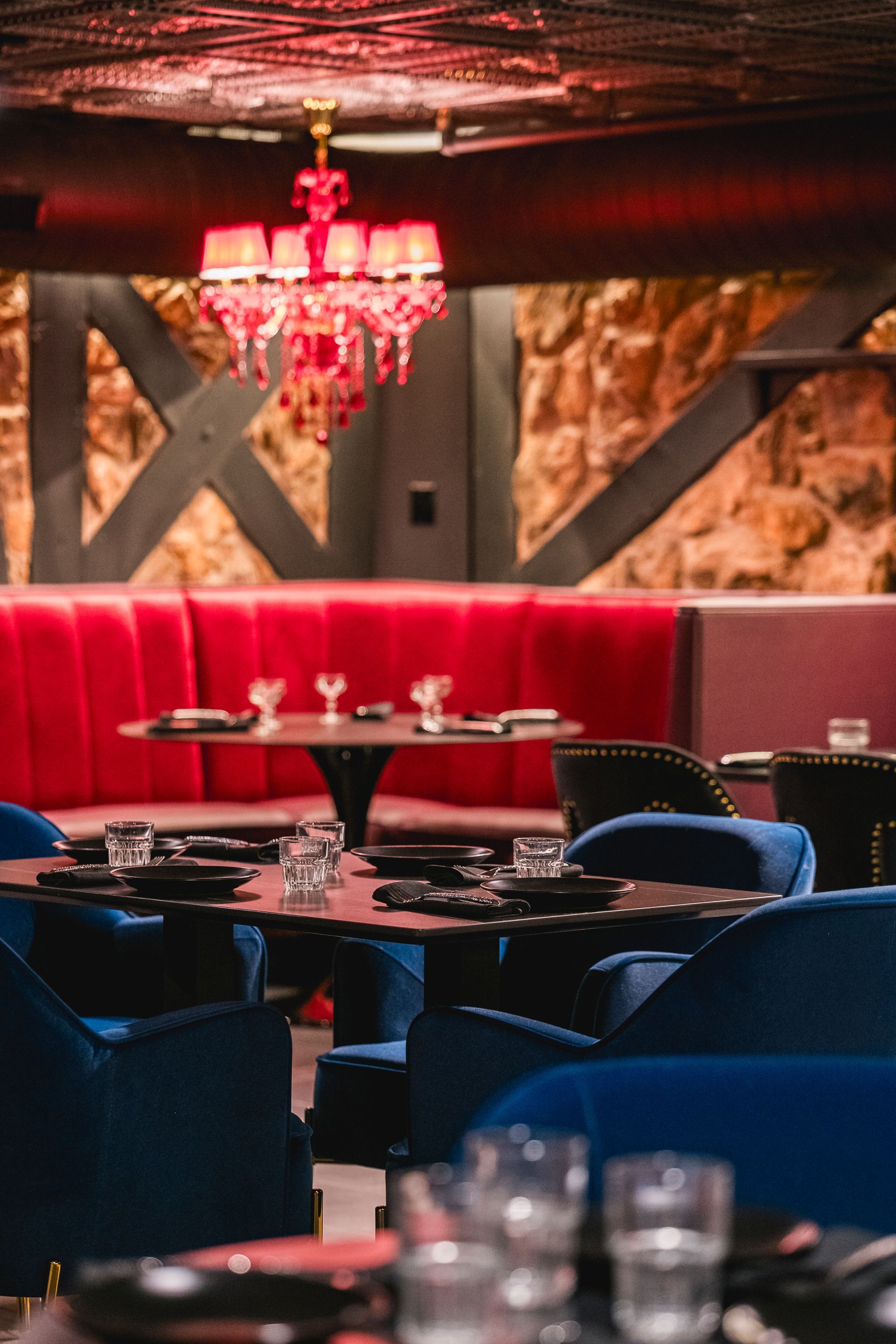 Dimly lit upscale lounge with red banquette seating, dark tables, blue chairs, and a red chandelier
