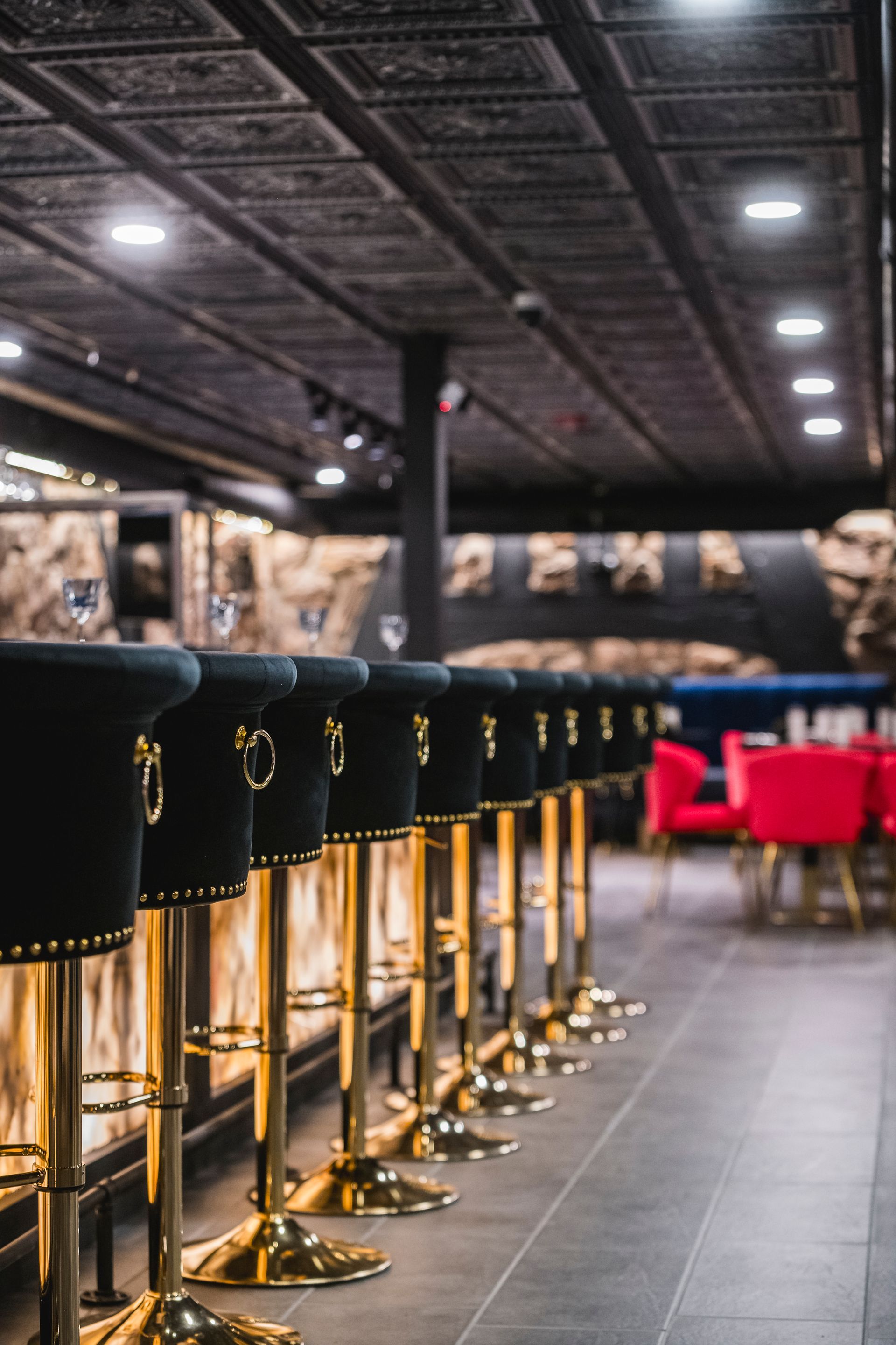 Empty bar with black stools, gold bases, and red chairs under a patterned ceiling.