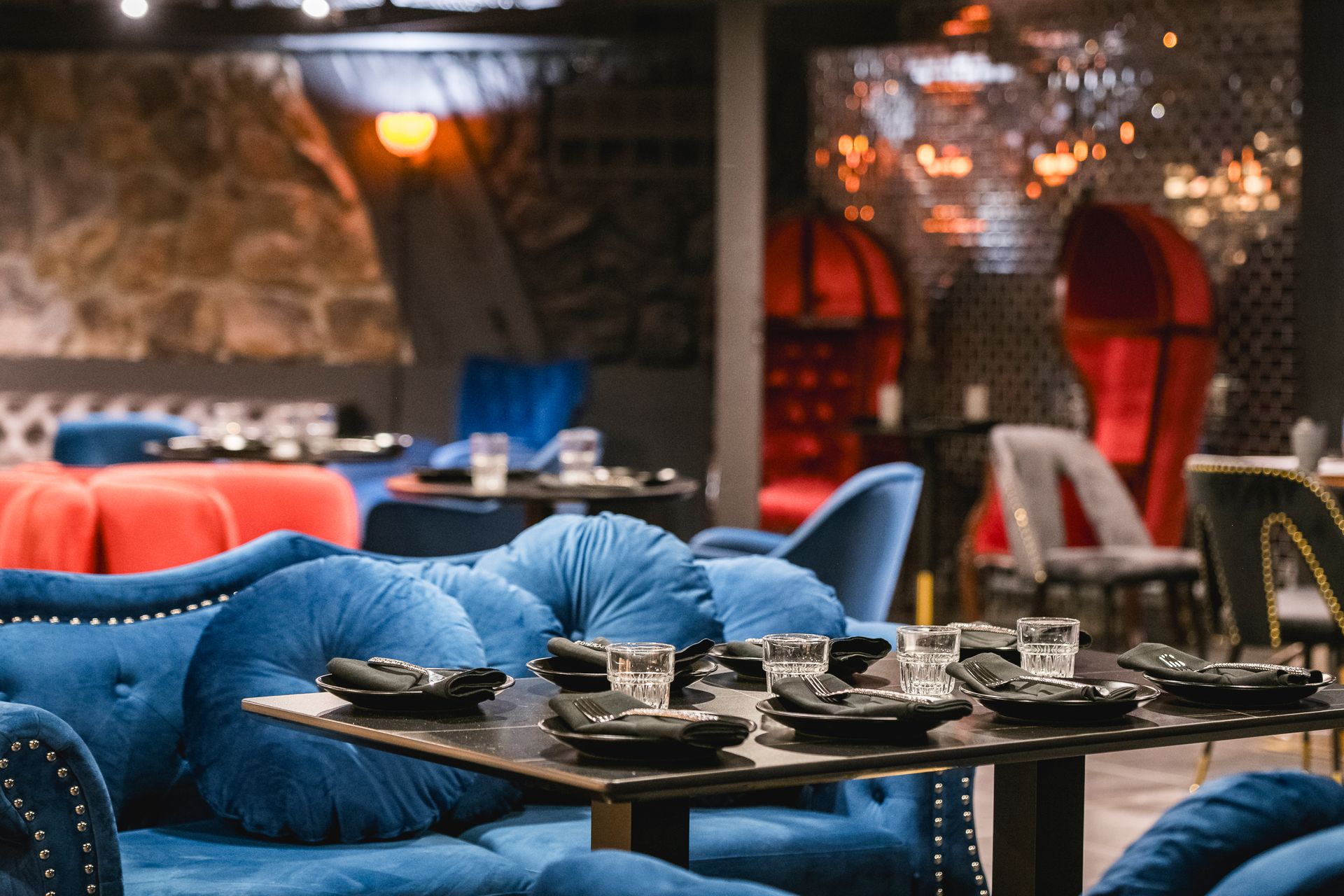Blue velvet lounge seating around dark tables in a dimly lit café or restaurant with red chairs in the background