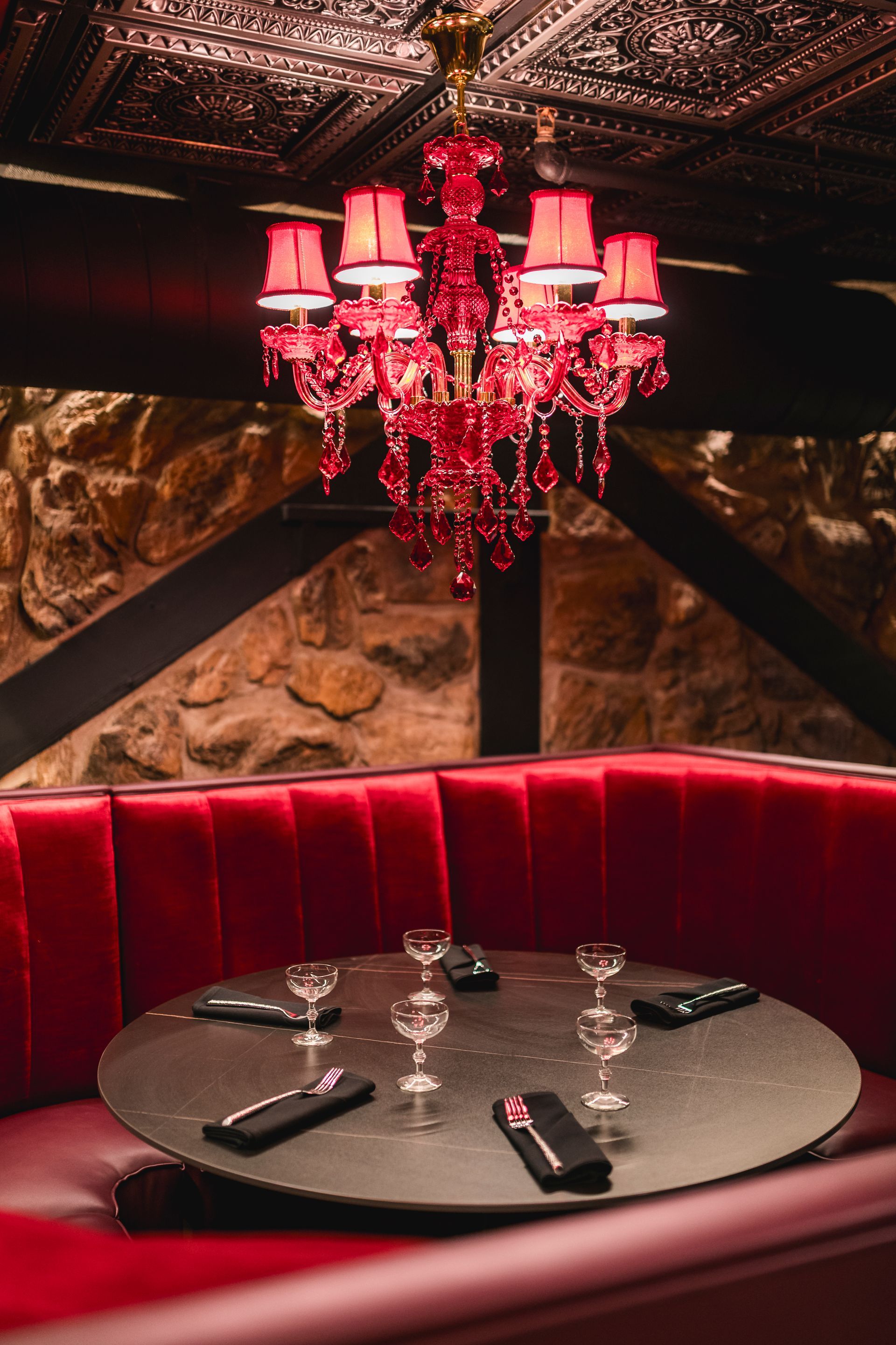 Dim red lounge booth with black table and pink chandelier under ornate ceiling