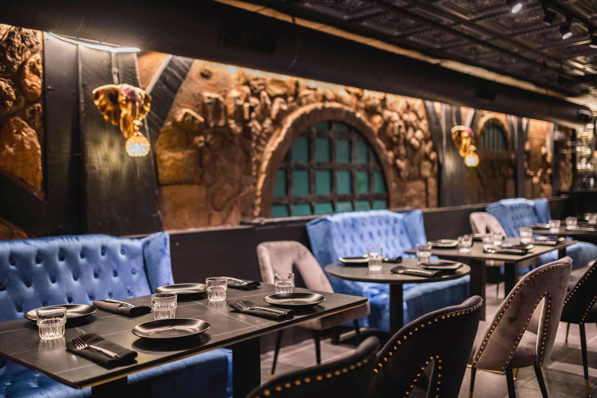 Elegant restaurant dining room with blue booths, set tables, and stone walls under warm pendant lights