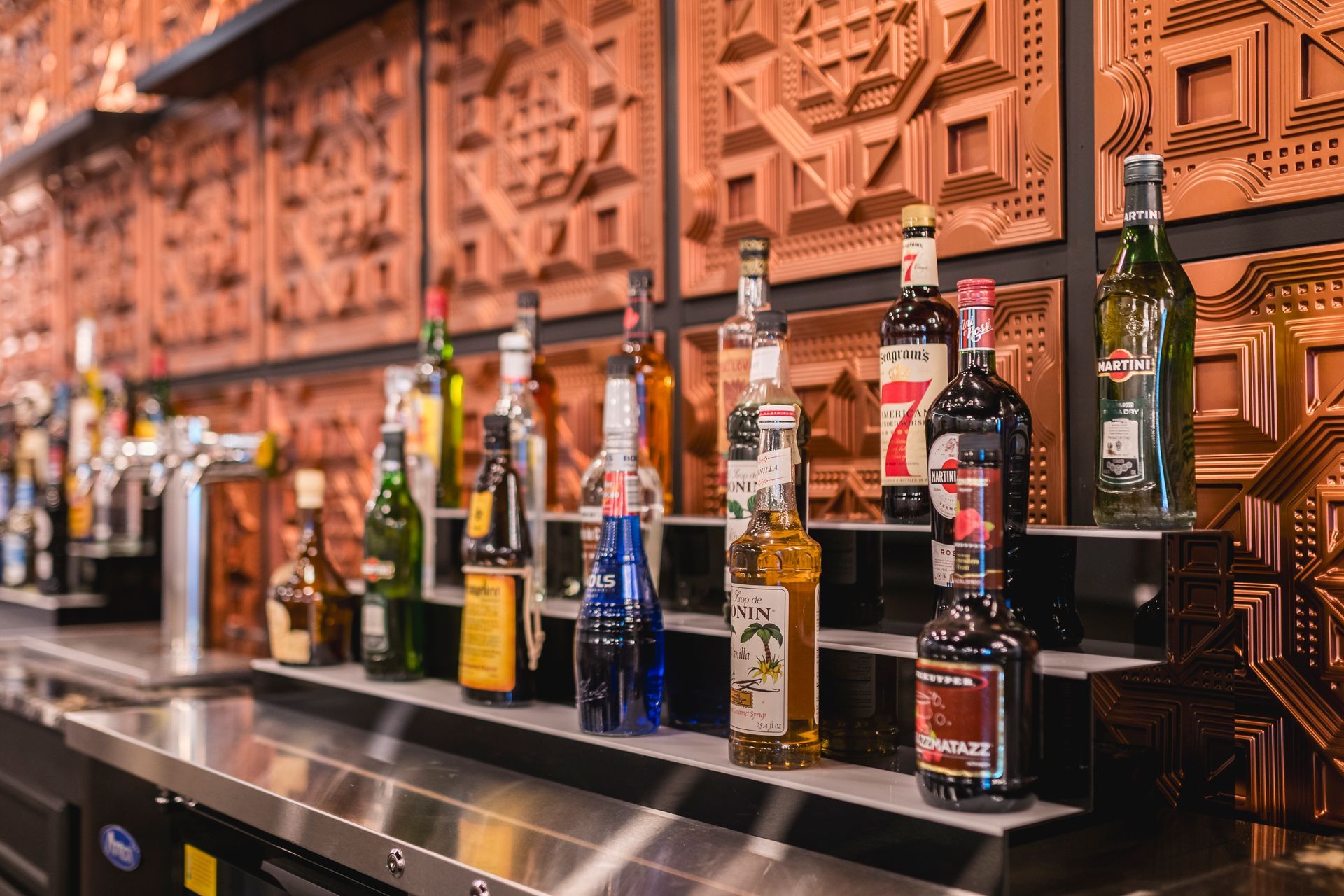 Row of liquor bottles on a bar counter with a copper-toned decorative wall behind them