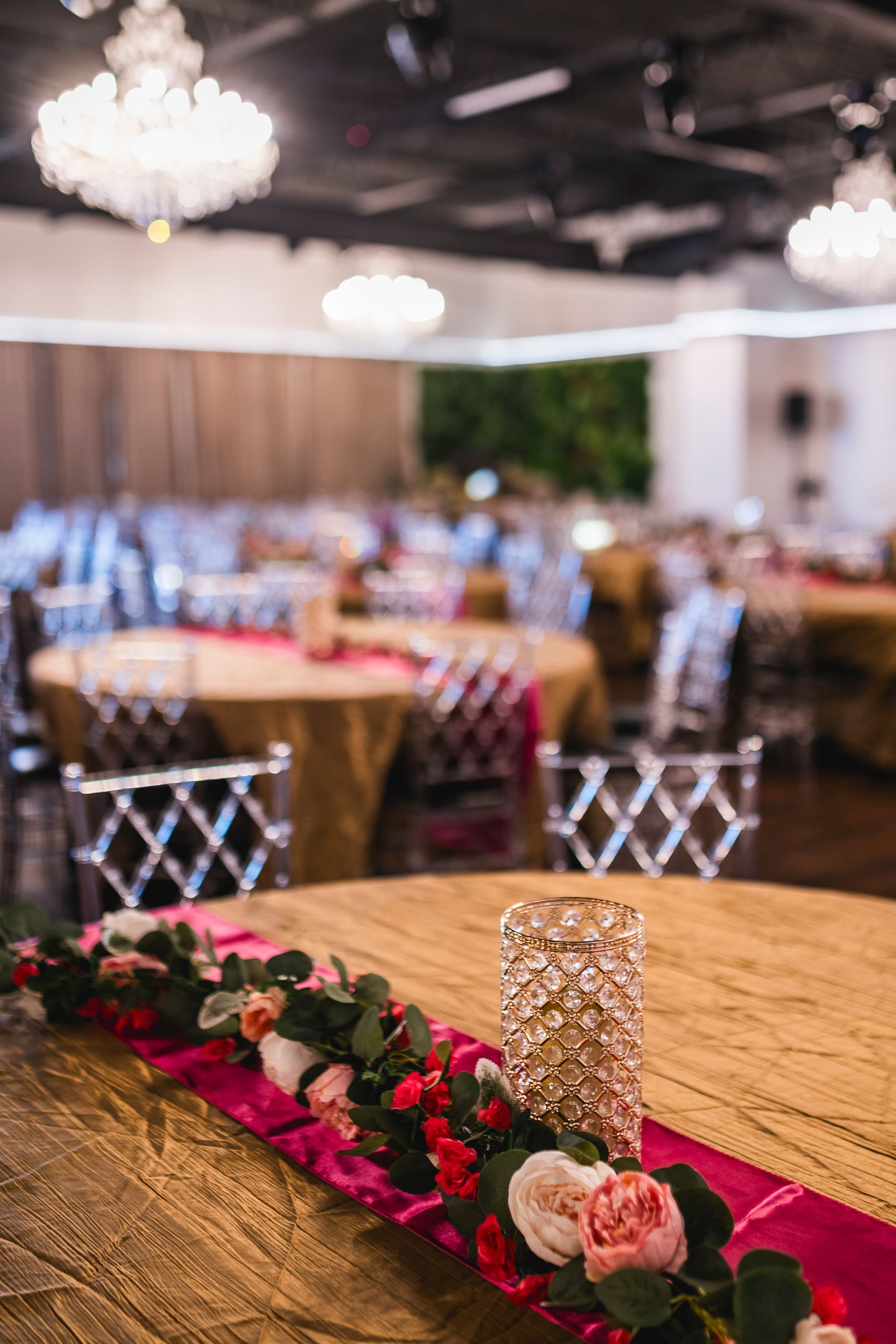 Close-up of a wooden banquet table with a pink runner, floral garland, and crystal candle holder in a decorated ballroom.