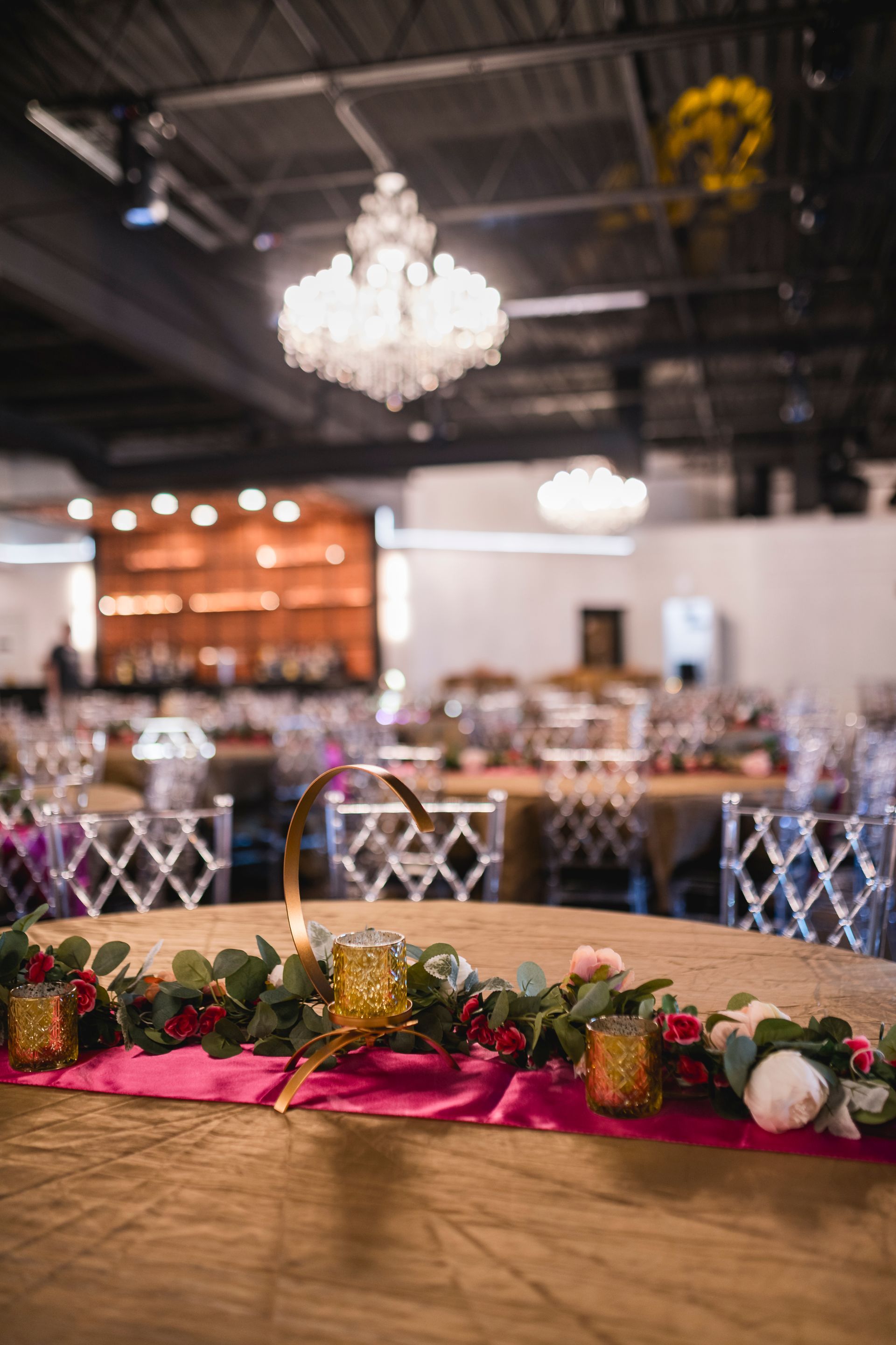 Banquet hall with chandelier, blurred dining tables, and a floral centerpiece on a gold-draped table in the foreground