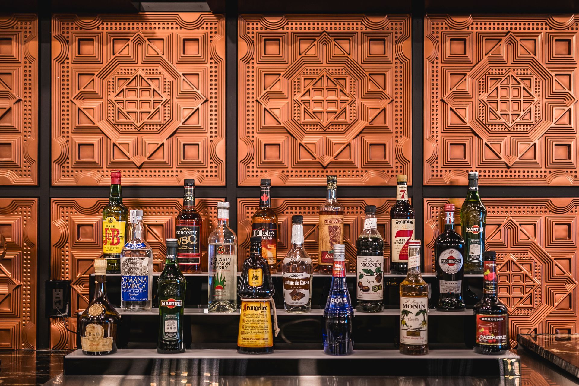 Bar counter lined with assorted liquor bottles beneath copper embossed wall panels.