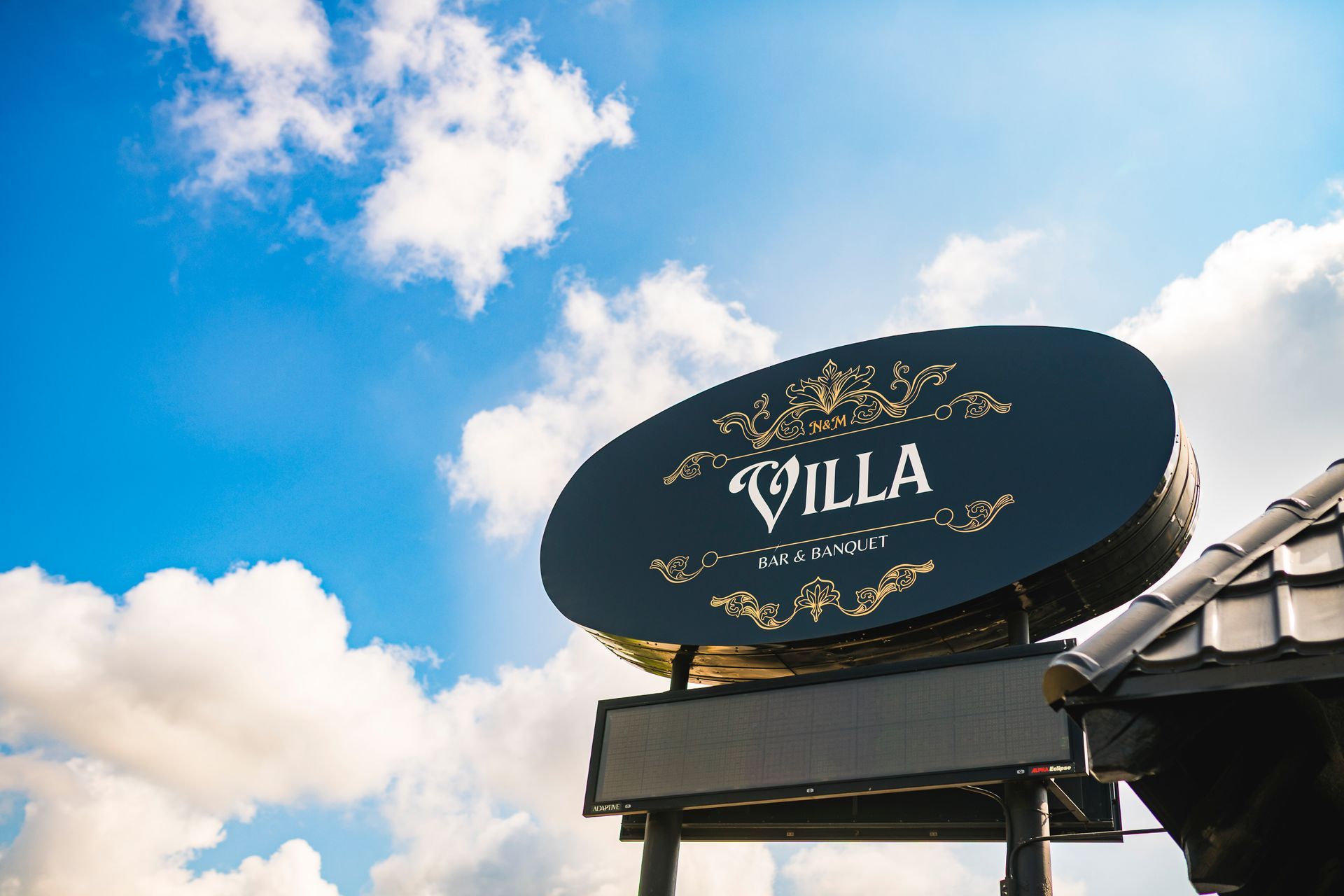 Oval black “VILLA” sign against a bright blue sky with clouds