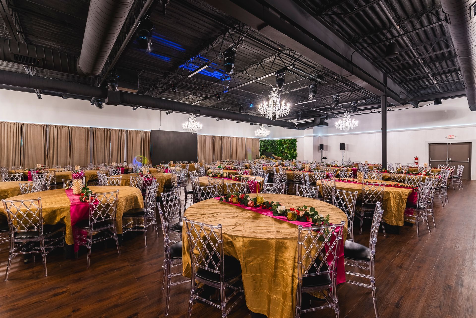 Banquet hall with round tables, gold tablecloths, red floral centerpieces, and chandeliers under a high black ceiling