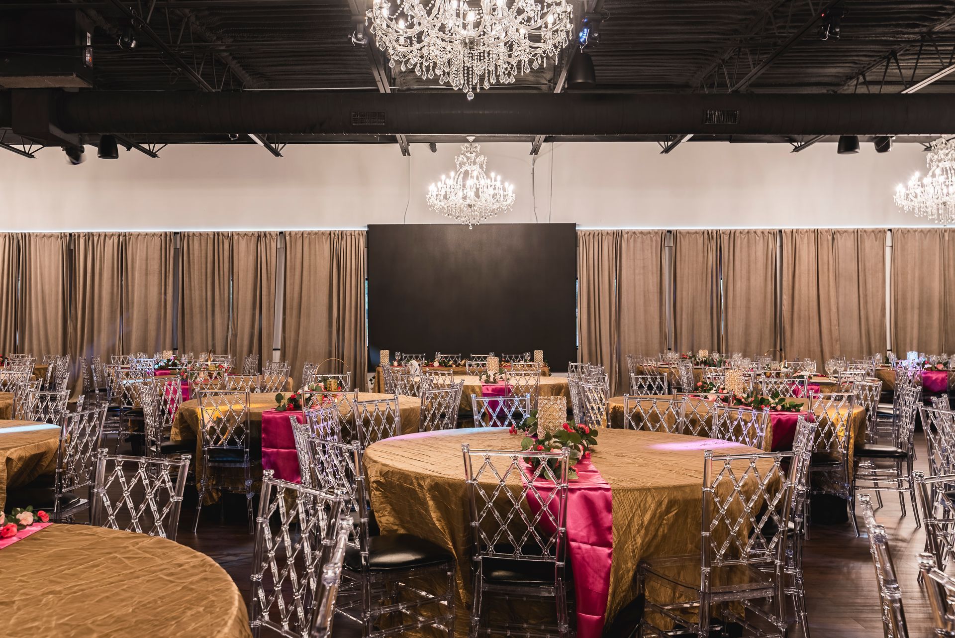 Elegant banquet hall with round tables, gold and pink linens, chandeliers, and clear chairs against draped curtains.