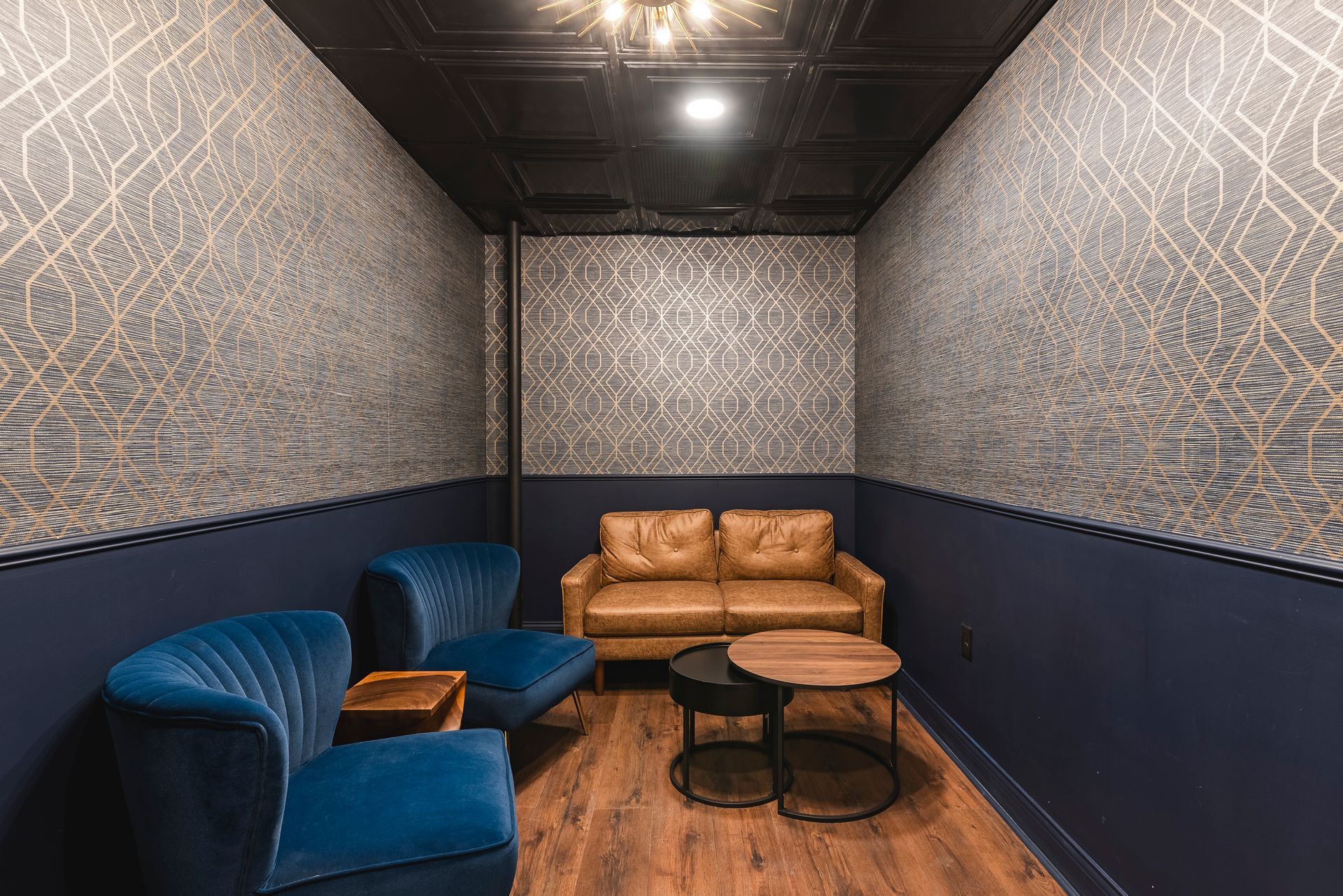 Small lounge with blue and tan sofas, round tables, patterned walls, and wooden floor under dim lighting