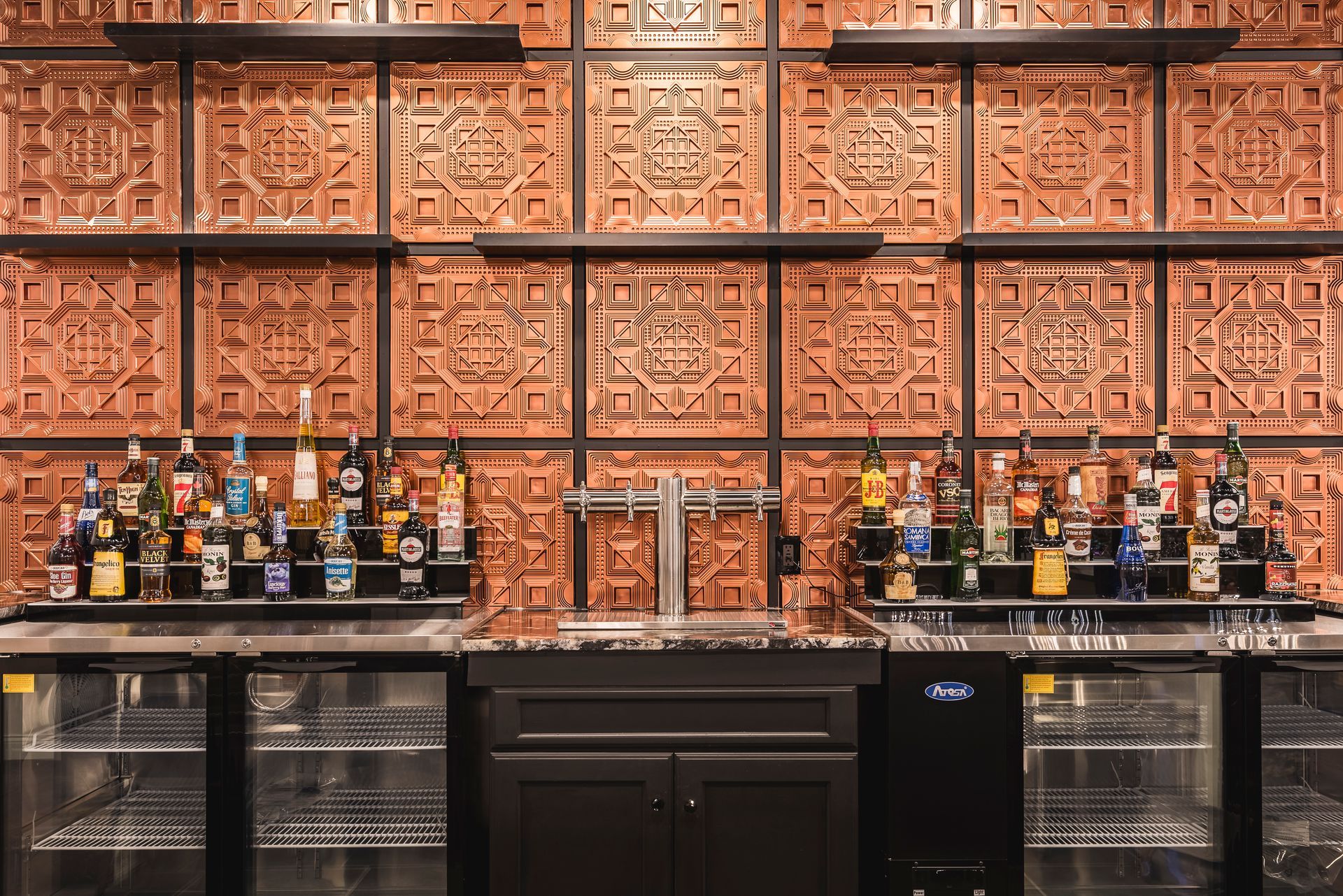 Bar shelves with assorted liquor bottles and glassware against an ornate copper-patterned wall