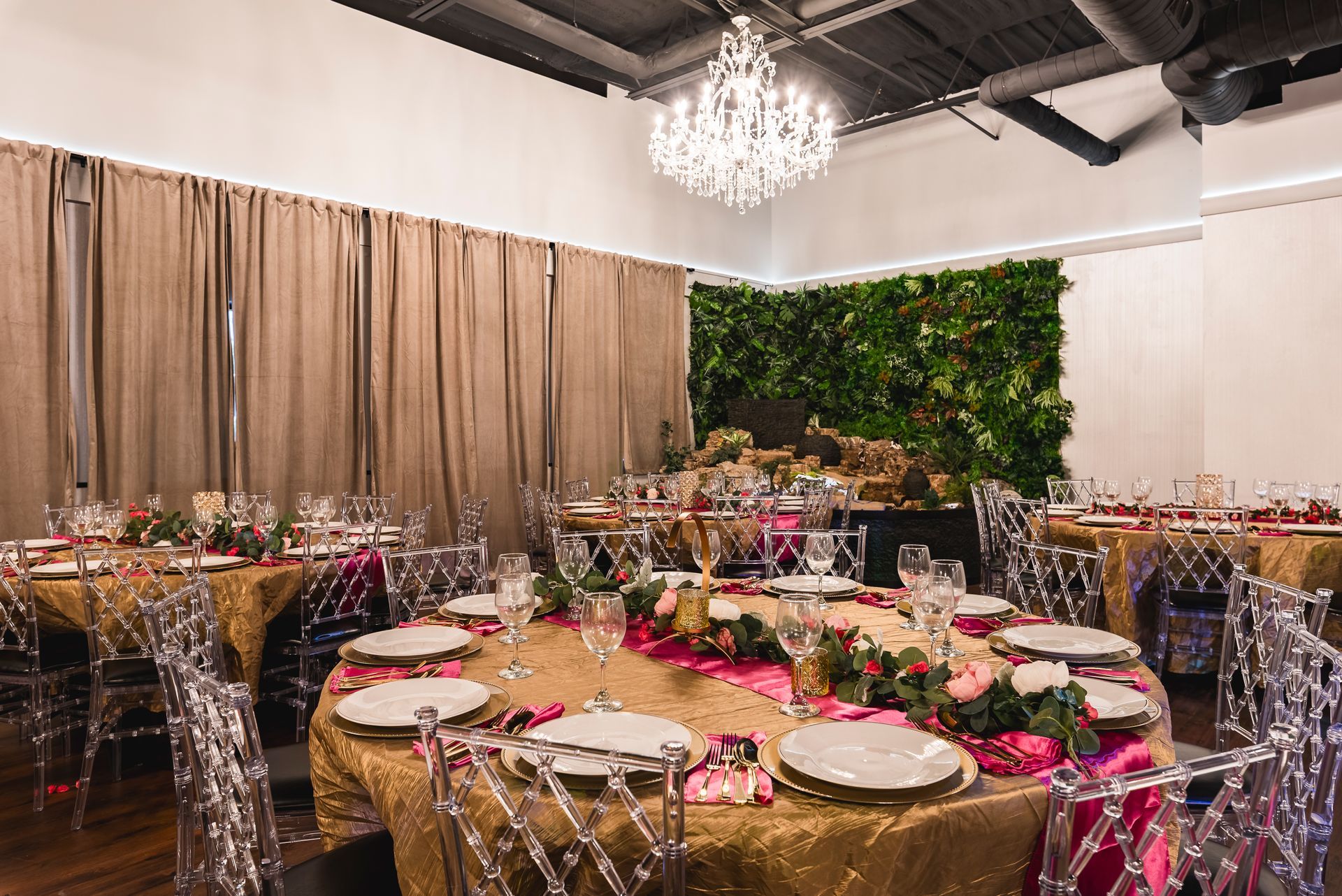 Elegant banquet hall with round tables, clear chairs, pink floral centerpieces, and a green wall backdrop.